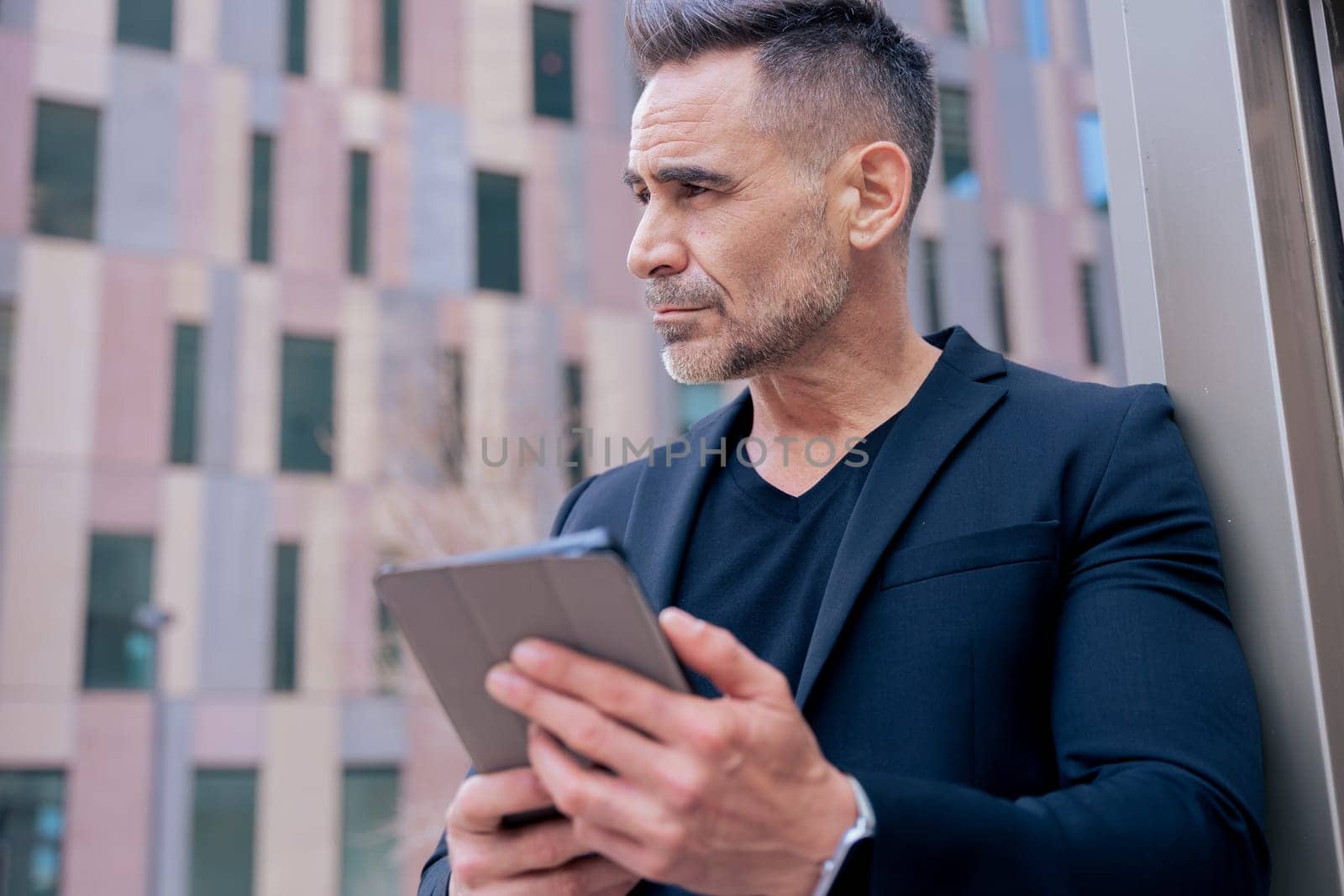 Businessman using a tablet outdoors in a modern city while looking away. He wears a black suit and smartwatch. Concept of technology, focus, mobility, and digital professional lifestyle.