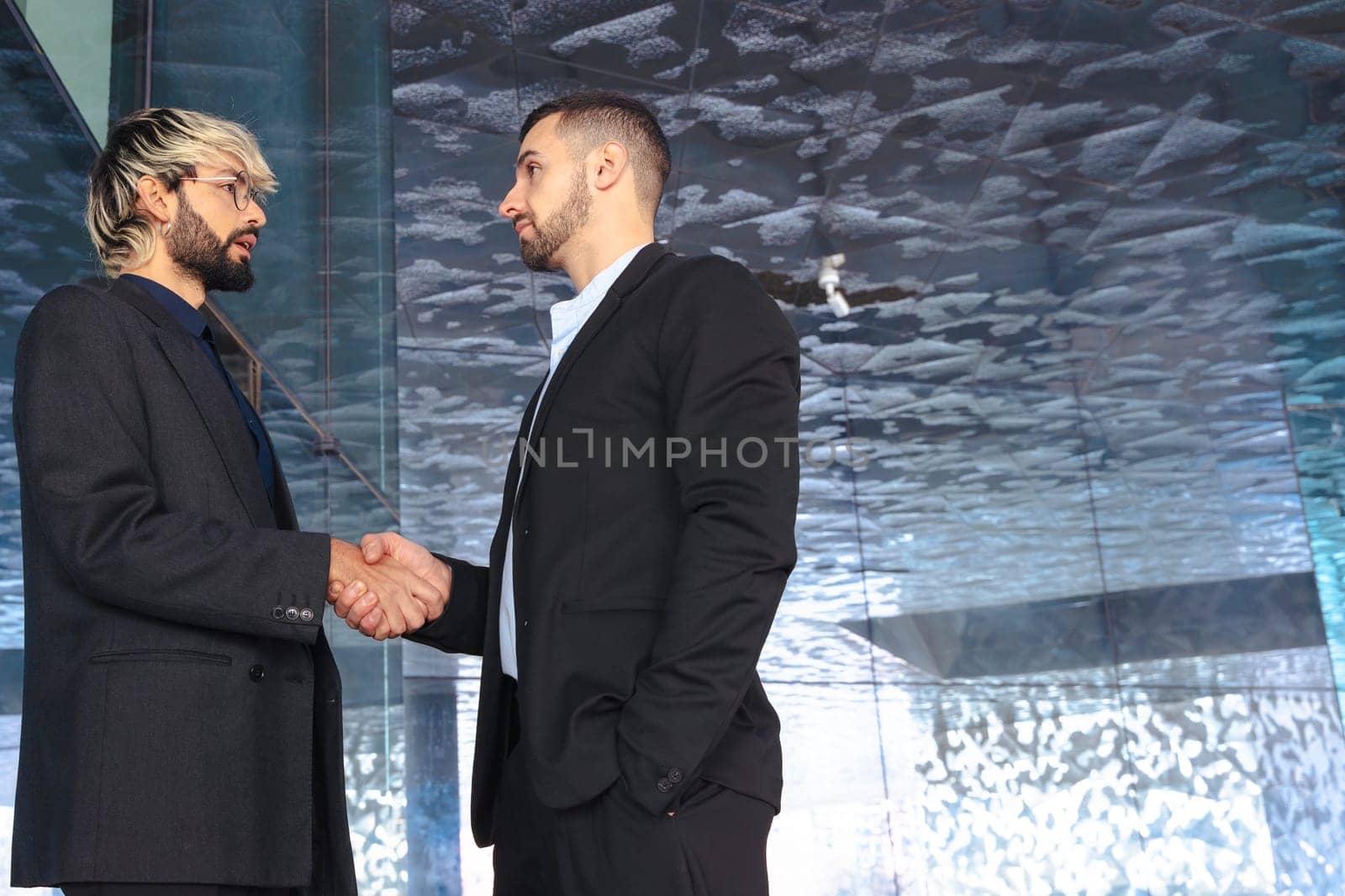 Two professional businessmen in suits shaking hands inside a modern glass office. Business deal, agreement, or partnership in a corporate indoor setting with contemporary design. Copy space