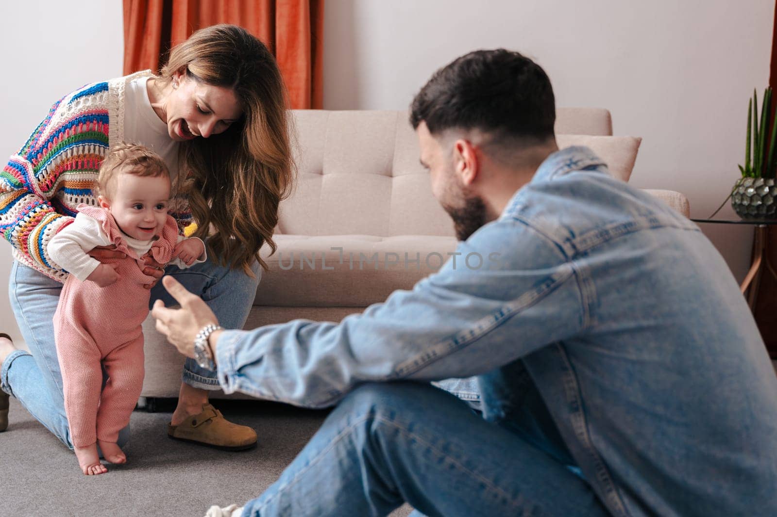 Mother and father encourage their baby girl as she takes her first steps on the carpet. A heartwarming family moment filled with love, support, and early childhood development at home.