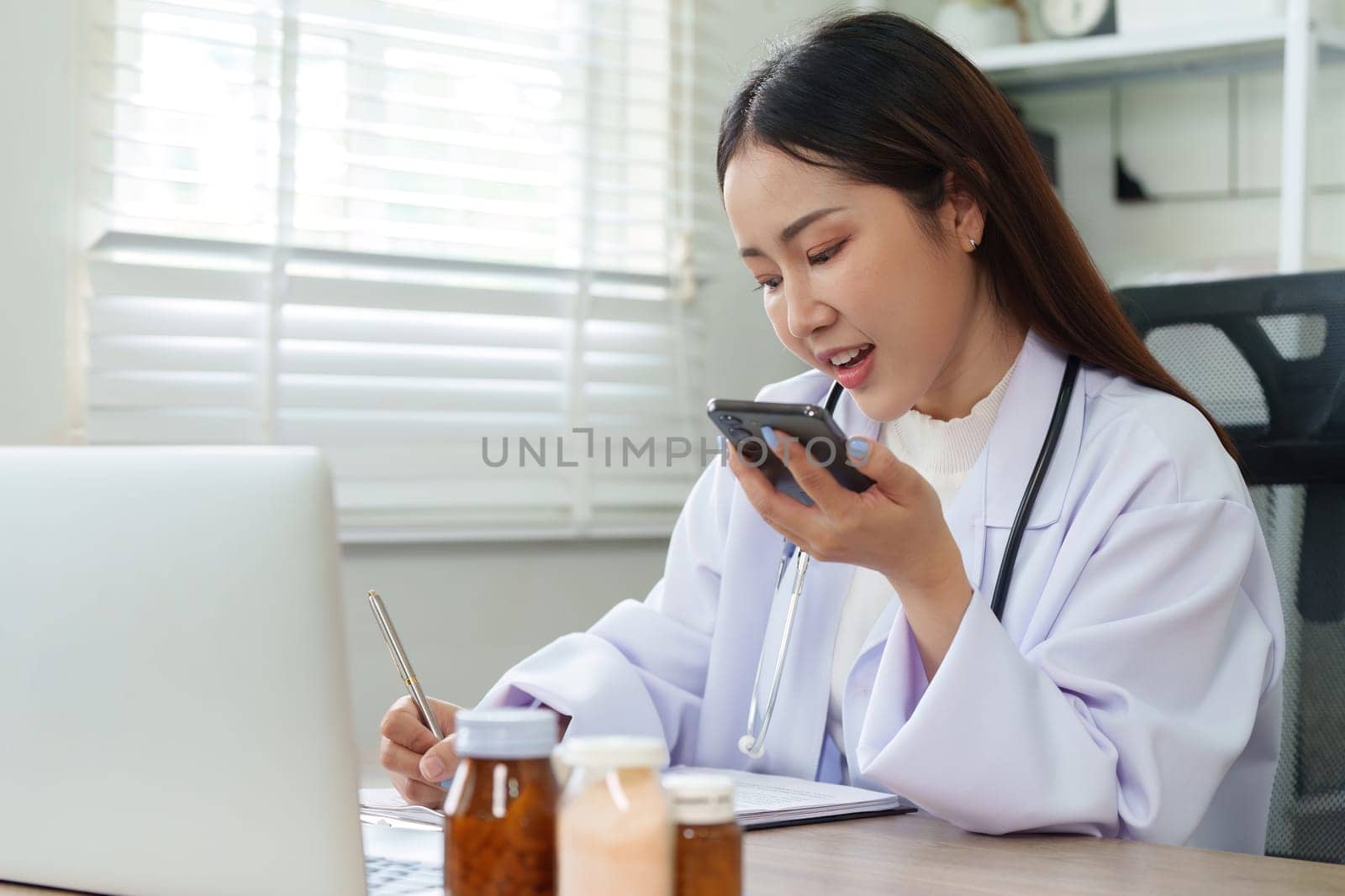 A female doctor engaged in a telehealth consultation, using a smartphone and laptop while taking notes, conveying professionalism and care.
