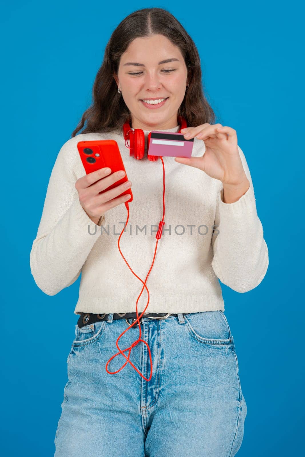 Happy young woman in a white sweater and red headphones smiling while showing a credit card to the camera and holding a red smartphone, against a blue background, symbolizing online shopping. Vertical
