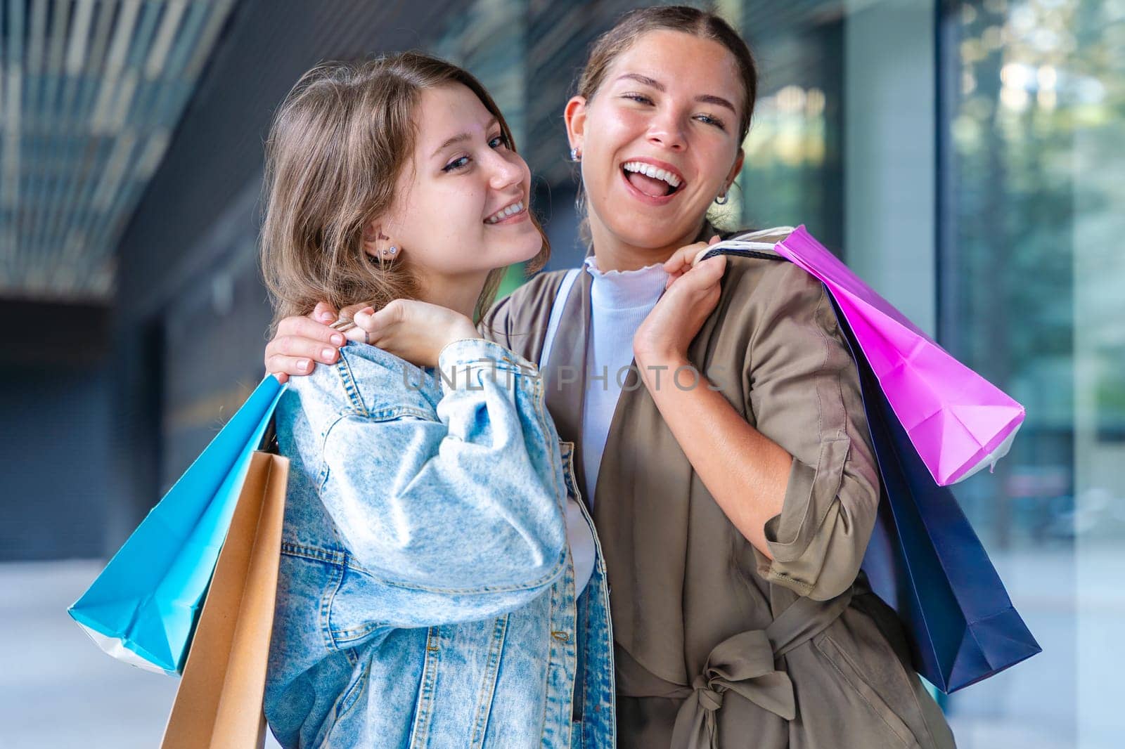 Two happy caucasian women holding colorful shopping bags and smiling after a successful day at the mall. They stand outside a modern building, enjoying their time together with style and joy.