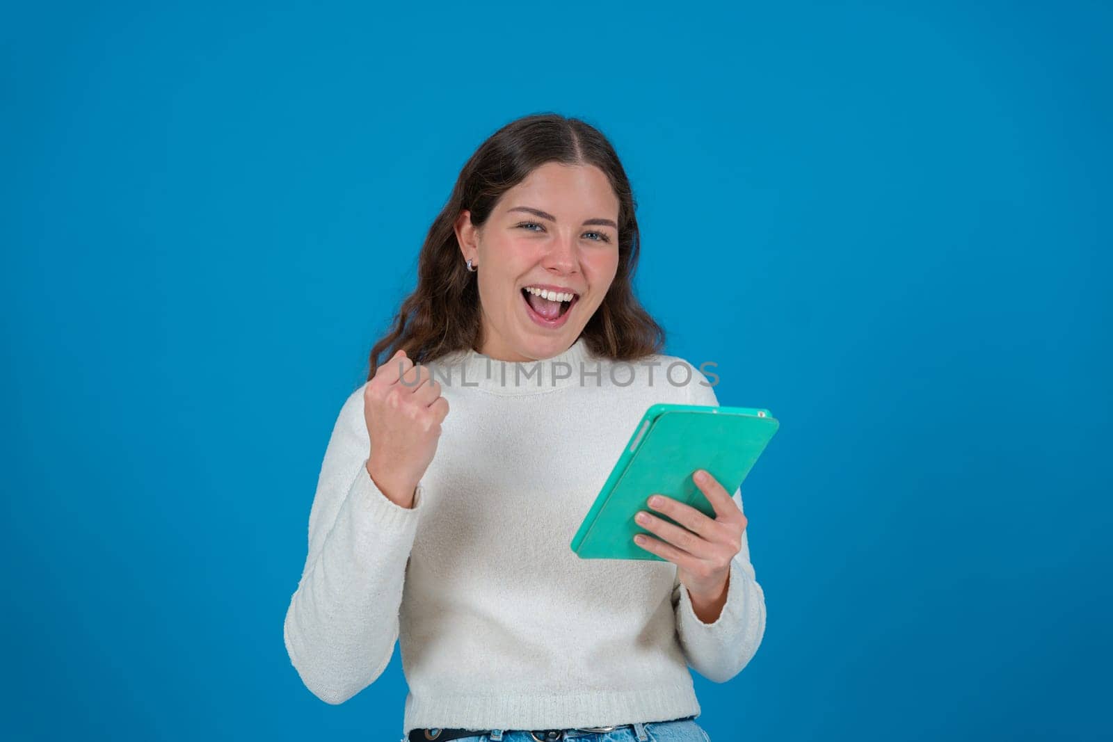 Happy young woman in a white sweater raising her fist in celebration while holding a green tablet and smiling at the camera, against a blue background, symbolizing success, joy, and digital life.