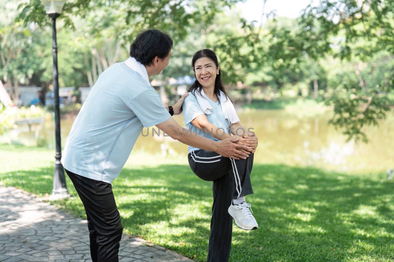 A senior man assists a woman with her stretching routine in a park, emphasizing the importance of wellness and fitness for seniors in a natural setting.