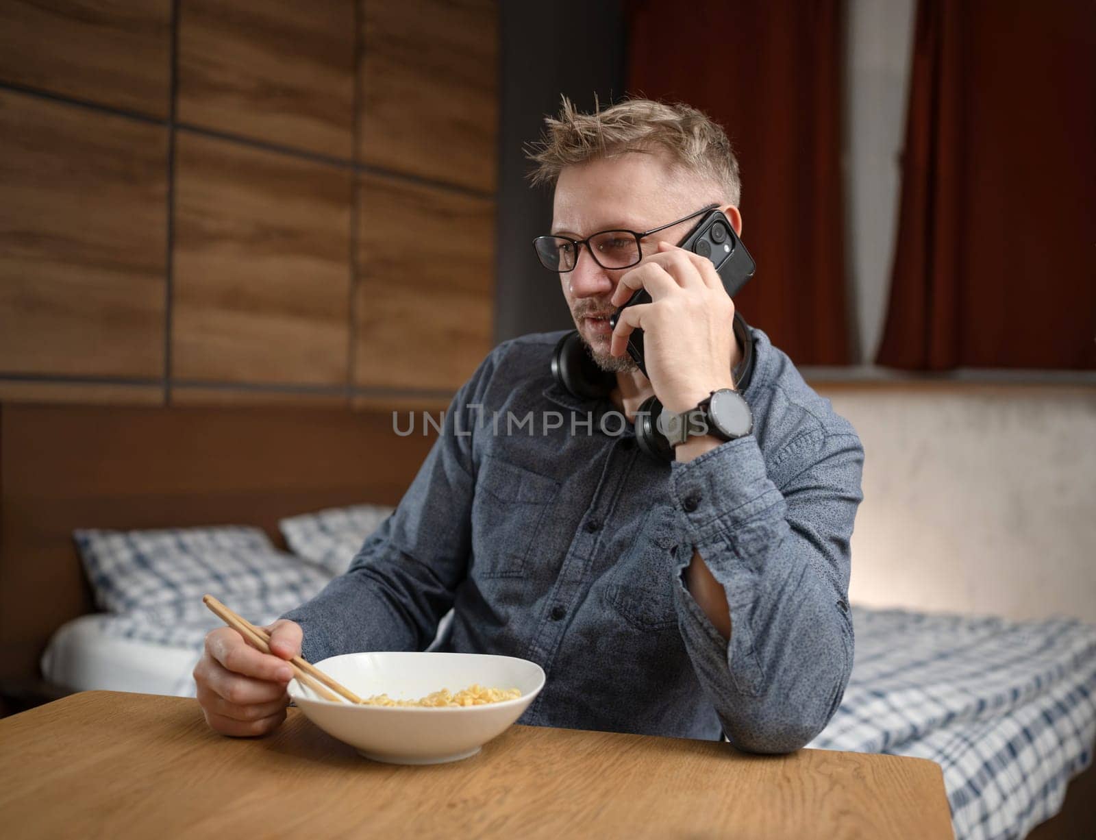 Handsome Man Eating Noodles With Chopsticks And Talking On The Phone At Home In The Bedroom