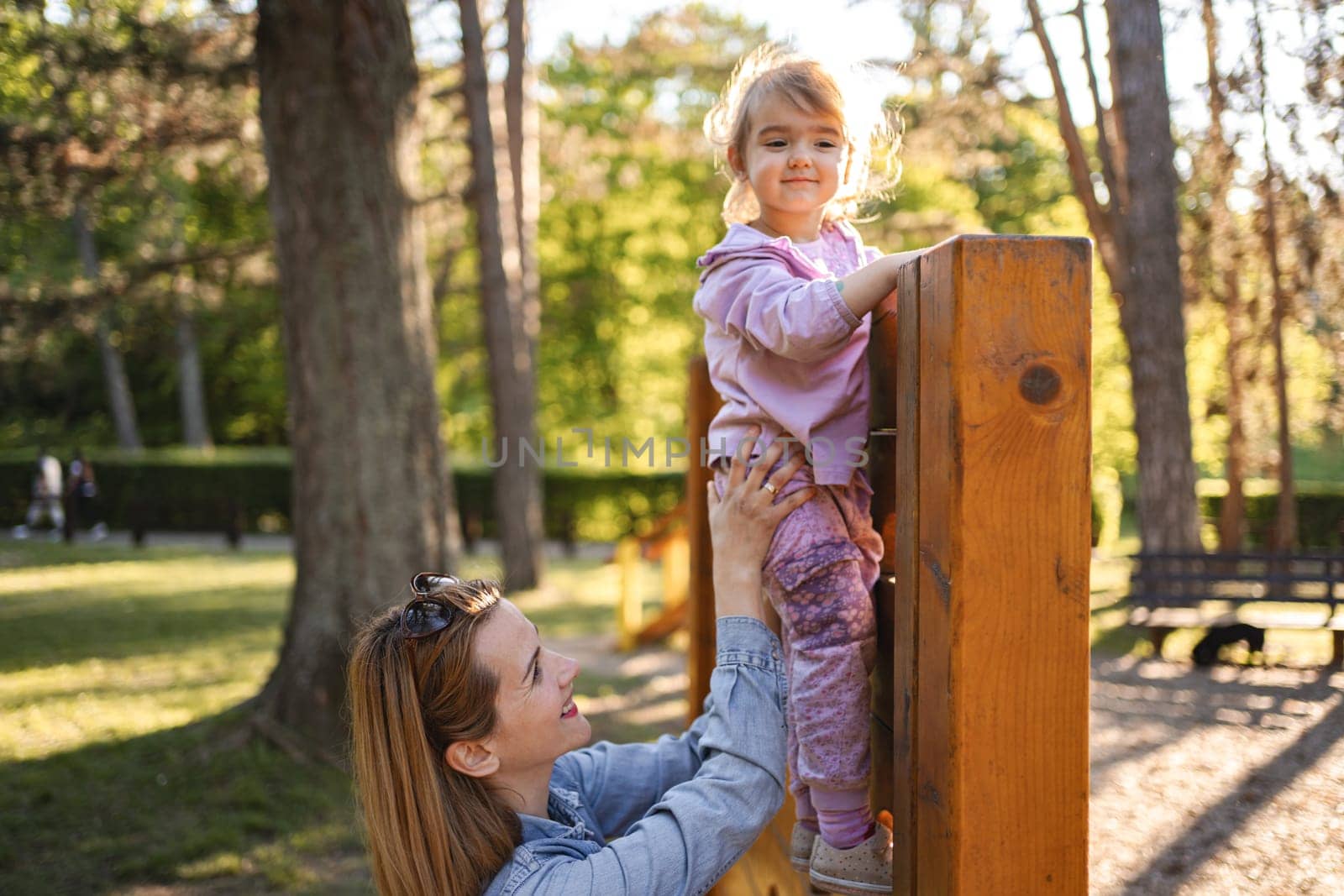 Smiling mom helps her cute daughter climb the playground.
