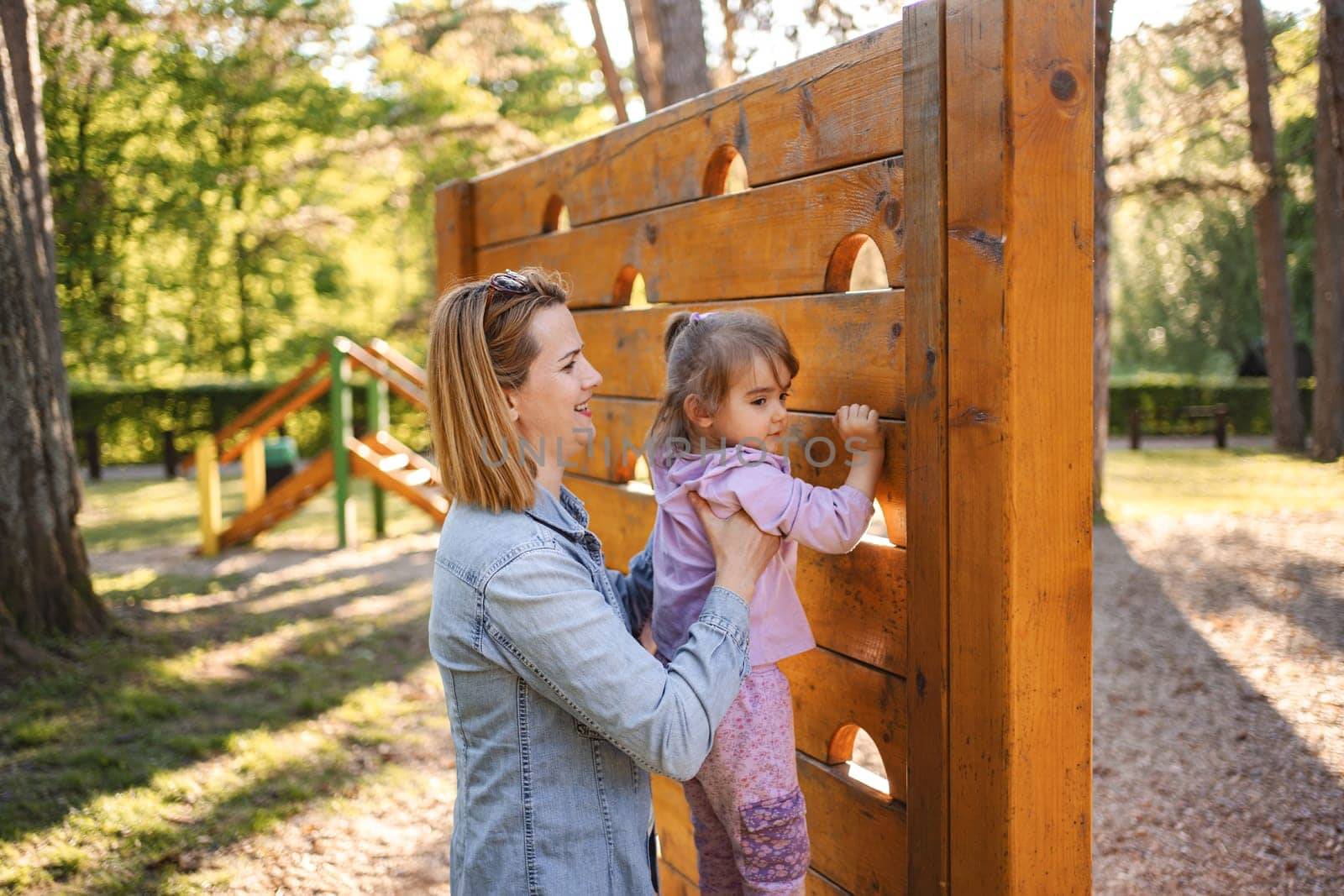 Smiling mom helps her cute daughter climb the playground.