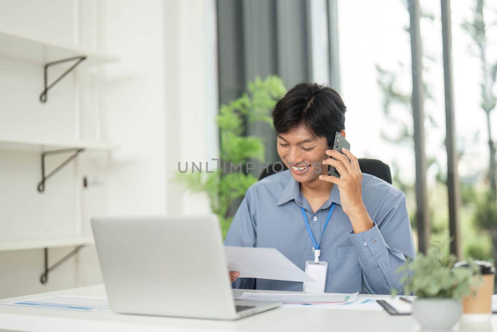 A cheerful young man talks on the phone while reviewing documents, emphasizing positive communication in a remote work setting.