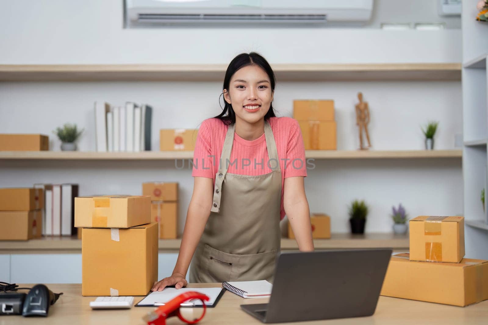A smiling young entrepreneur stands confidently with packages in her home office, representing small business success and positivity.
