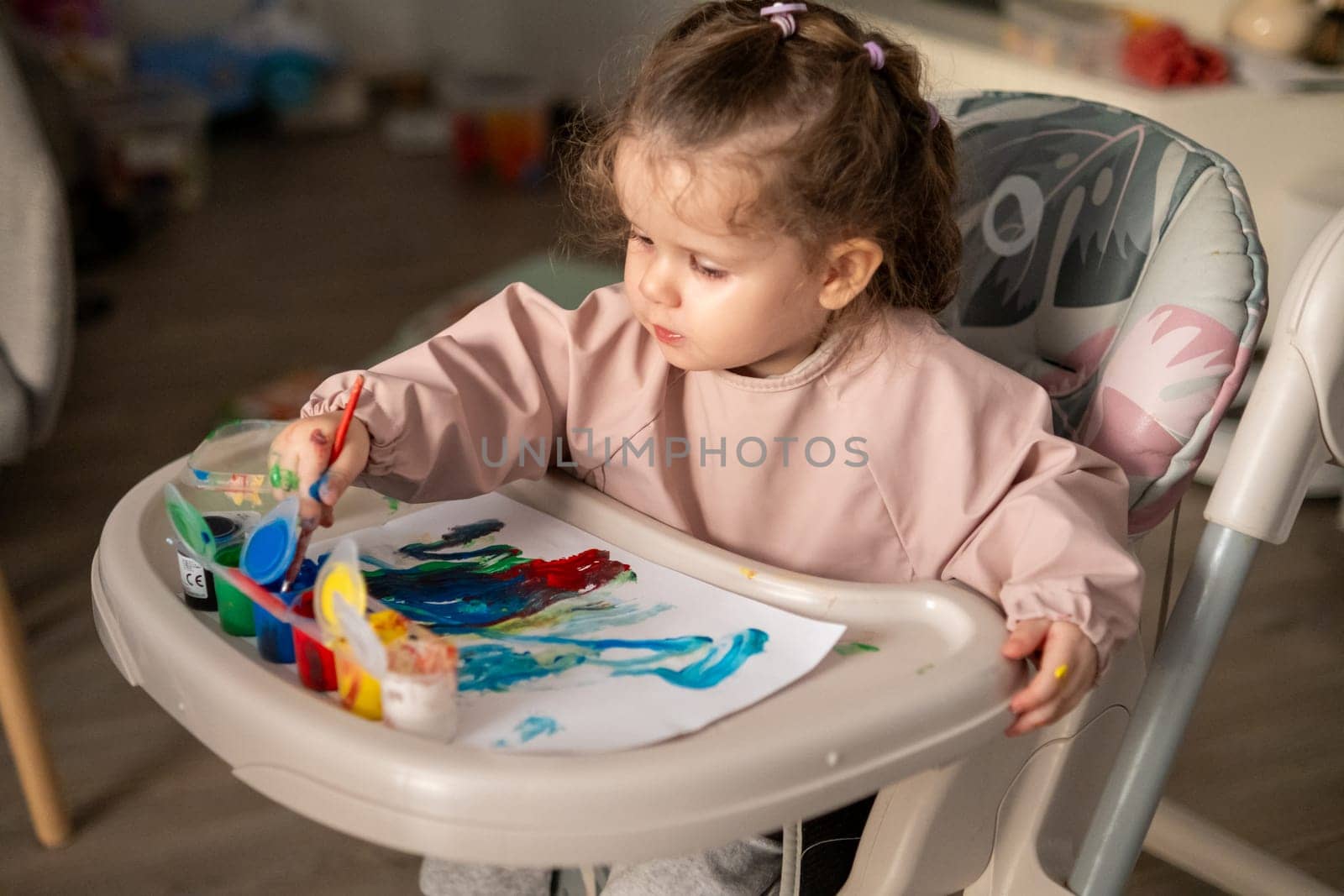 Young girl painting with gouache at a kids table, surrounded by toys and open paint jars. Artistic mess and imaginative play.