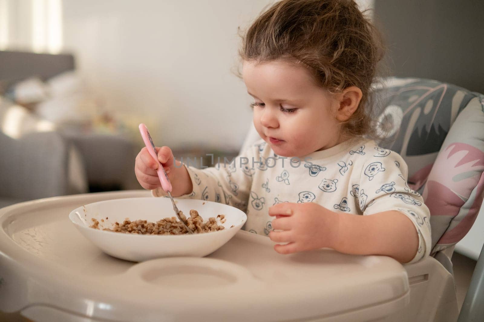 Cute little girl with curly hair sitting in a baby chair, enjoying oatmeal in a bright kitchen. Childhood mealtime moments.
