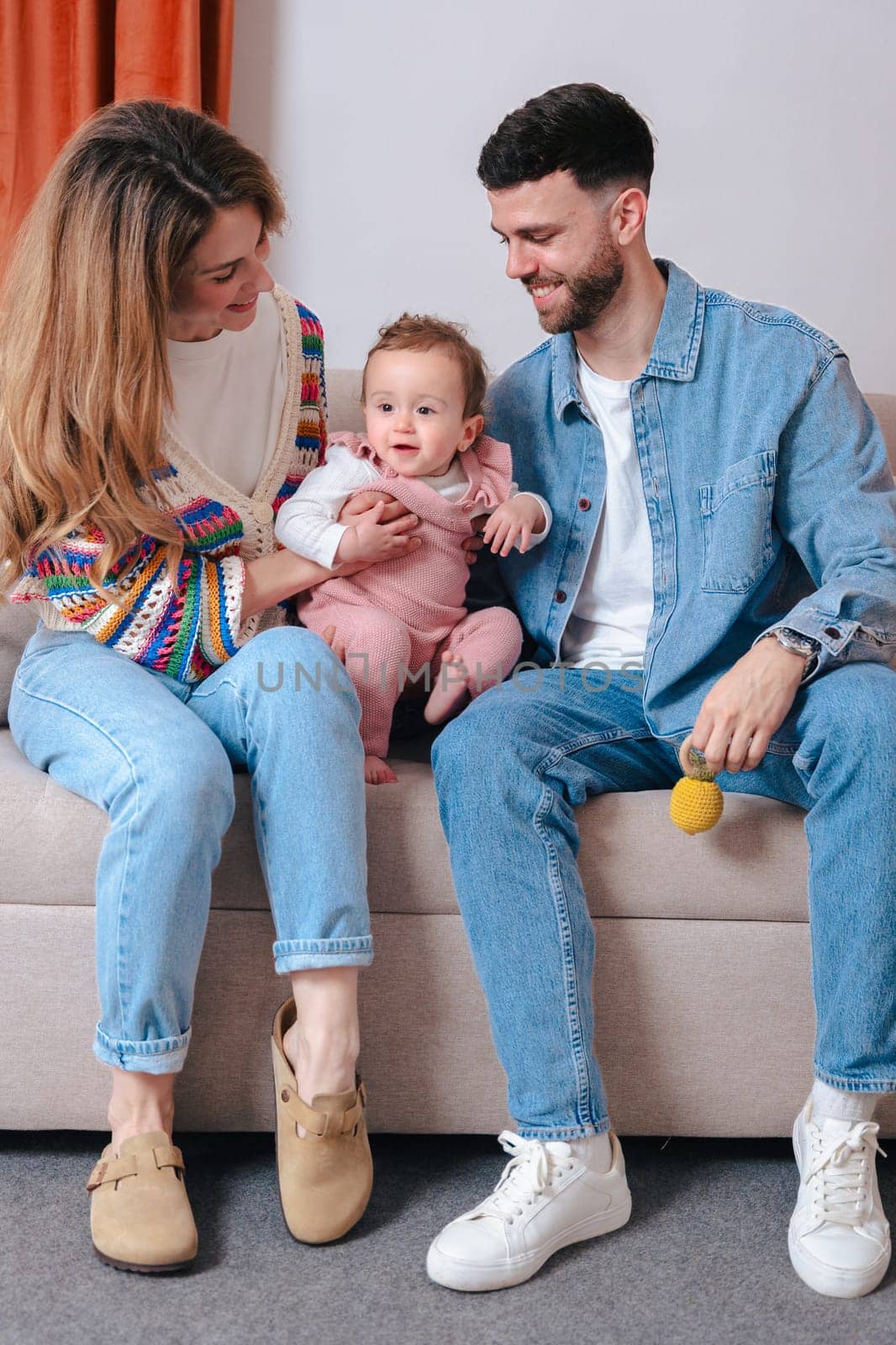 Vertical portrait Smiling parents enjoy playtime with their baby girl who holds toys while sitting on a sofa. A joyful family bonding moment filled with love, color, and warmth in a cozy home setting.