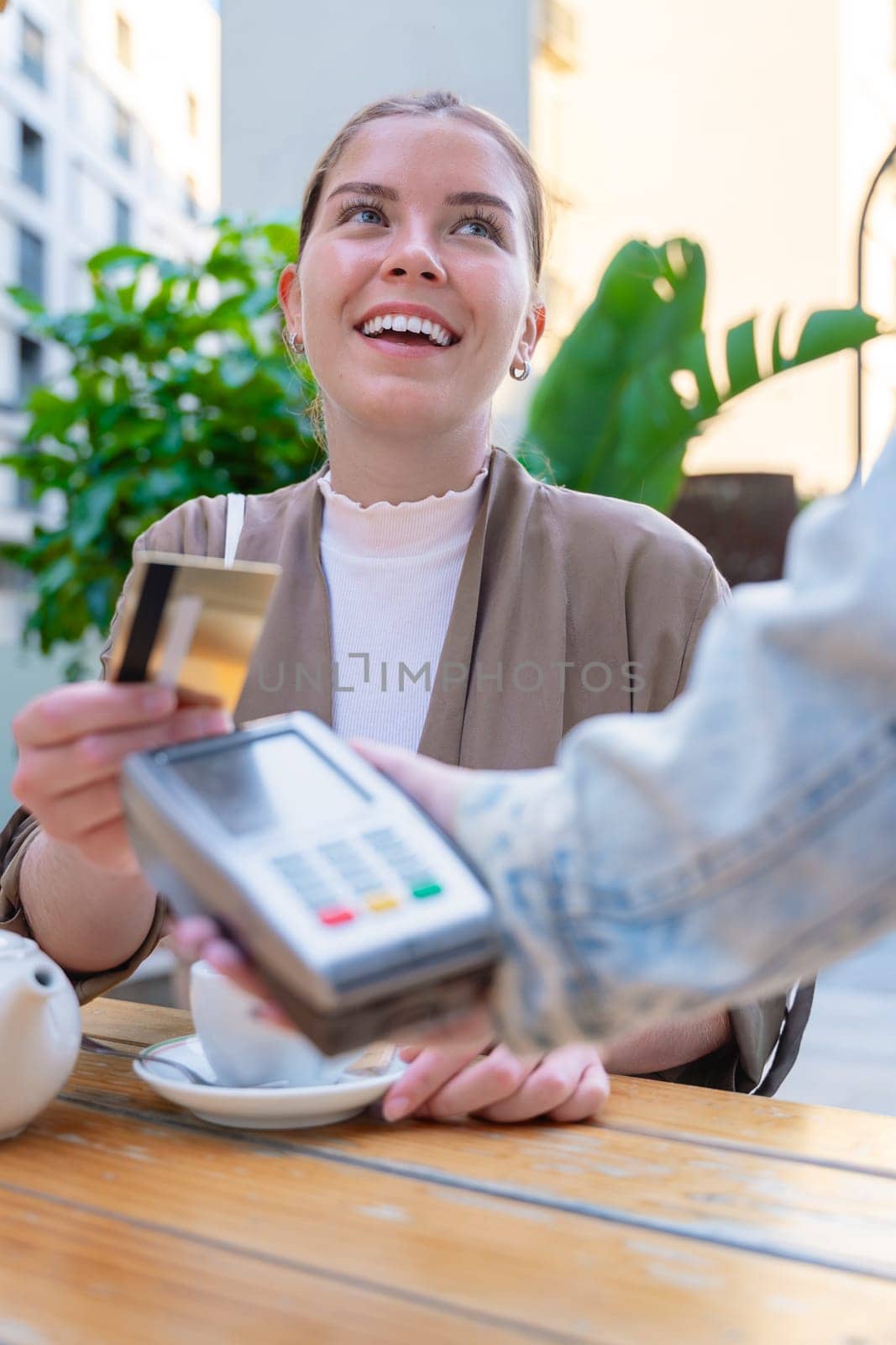 Caucasian woman smiling as she pays with a credit card using contactless technology at an outdoor cafe. A casual urban moment reflecting convenience, lifestyle, and modern financial habits. Vertical