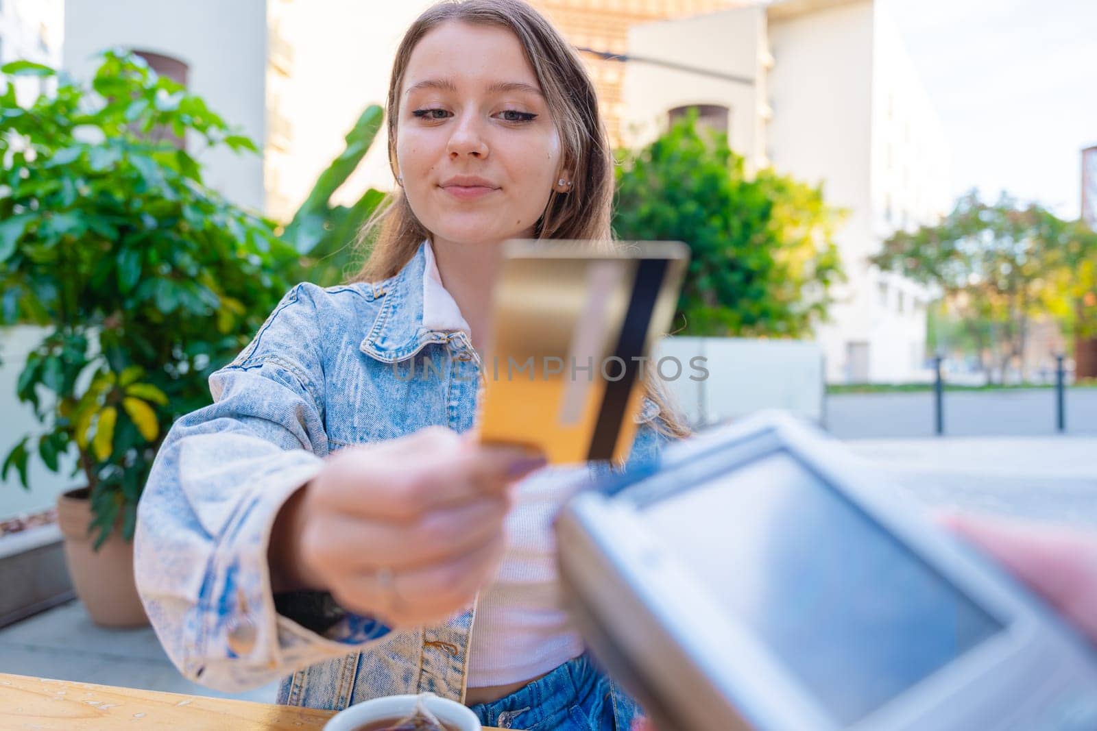 Caucasian woman smiling as she pays with a credit card using contactless technology at an outdoor cafe. A casual urban moment reflecting convenience, lifestyle, and modern financial habits.