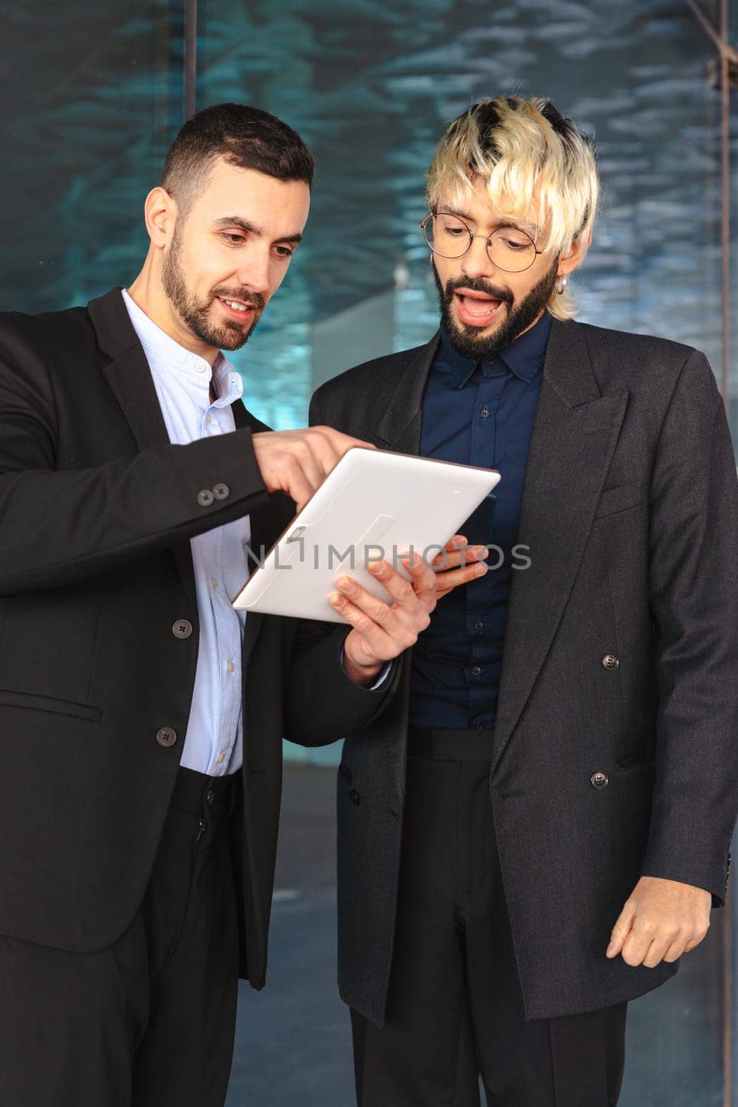 Two young businessmen in formal suits ,looking at a tablet and smiling while working together in a stylish modern office with reflective surfaces and a professional atmosphere. Vertical