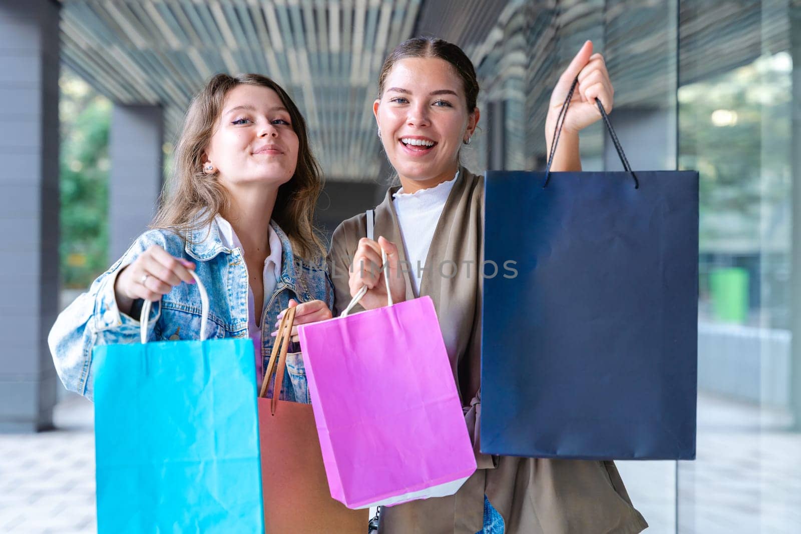 Two cheerful Caucasian women looking at the camera and smiling while holding colorful shopping bags. Capturing joyful and fun shopping moments in an urban outdoor setting.