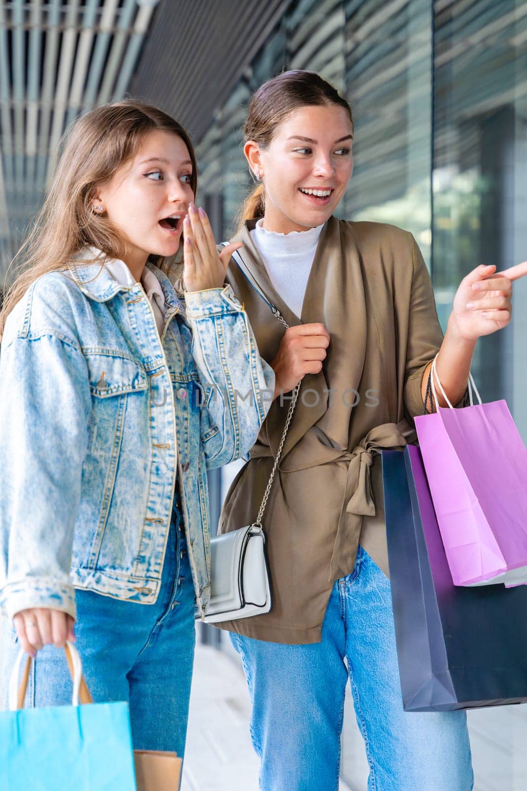 Two happy Caucasian women holding shopping bags and pointing at a store window display. Candid and joyful urban moment during a day of shopping and friendship in the city. Vertical