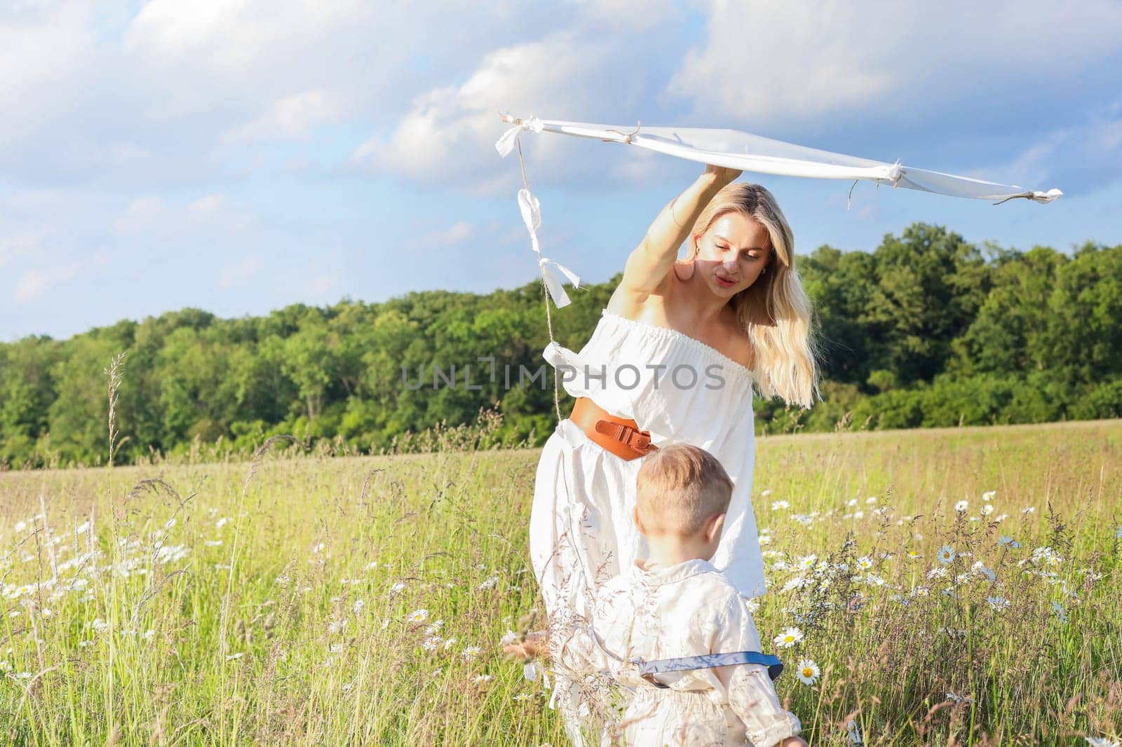 In a meadow in the grass and wildflowers, a child and a mother in a white dress and with blond hair are launching a kite into the sky, having fun with their little son. High quality photo