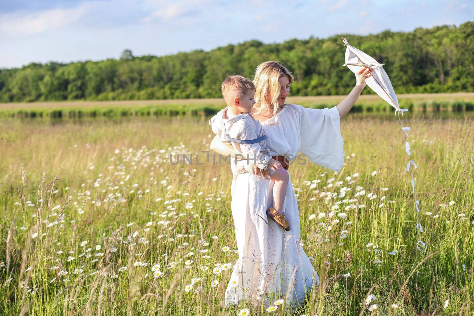 In a meadow in the grass and wildflowers, a child and a mother in a white dress and with blond hair are launching a kite into the sky, having fun with their little son. High quality photo