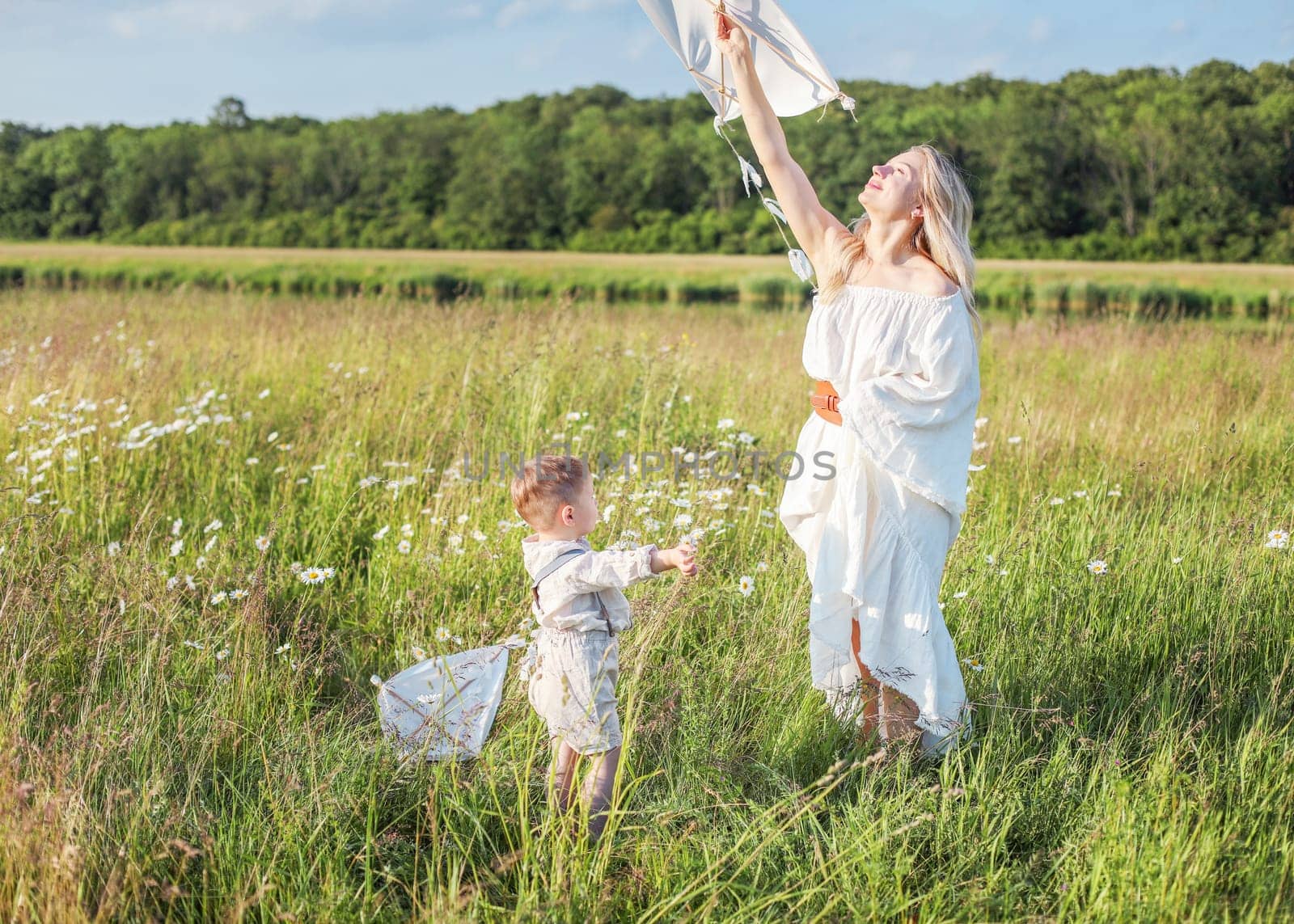 In a meadow in the grass and wildflowers, a child and a mother in a white dress and with blond hair are launching a kite into the sky, having fun with their little son. High quality photo