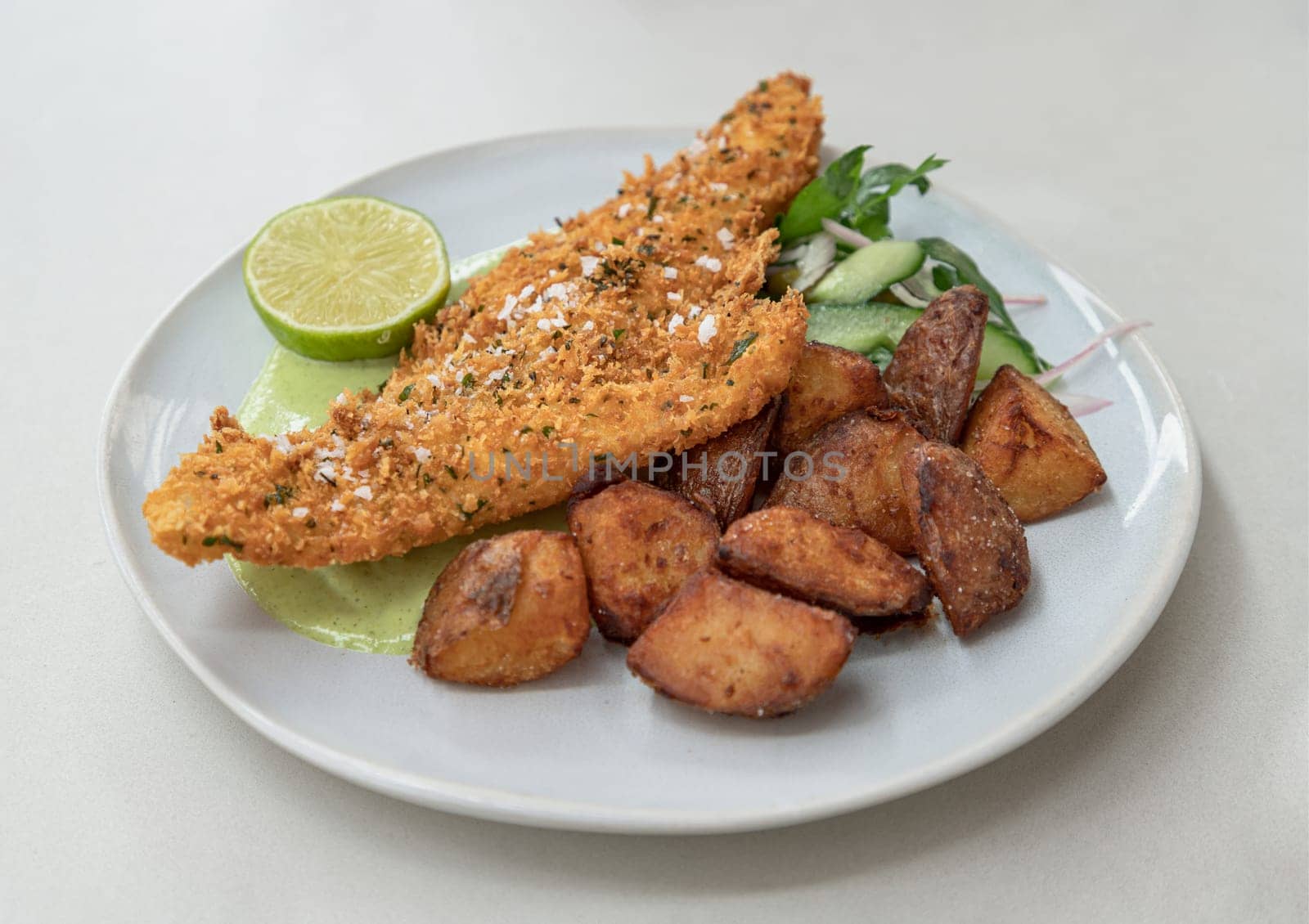 Homemade Fish and chips served with Lime cut in half, Cucumber slices, Rocket, Shallots and Green tartar sauce on Ceramic plate. use it as your Wallpaper, Poster and Copy space, Selective focus.