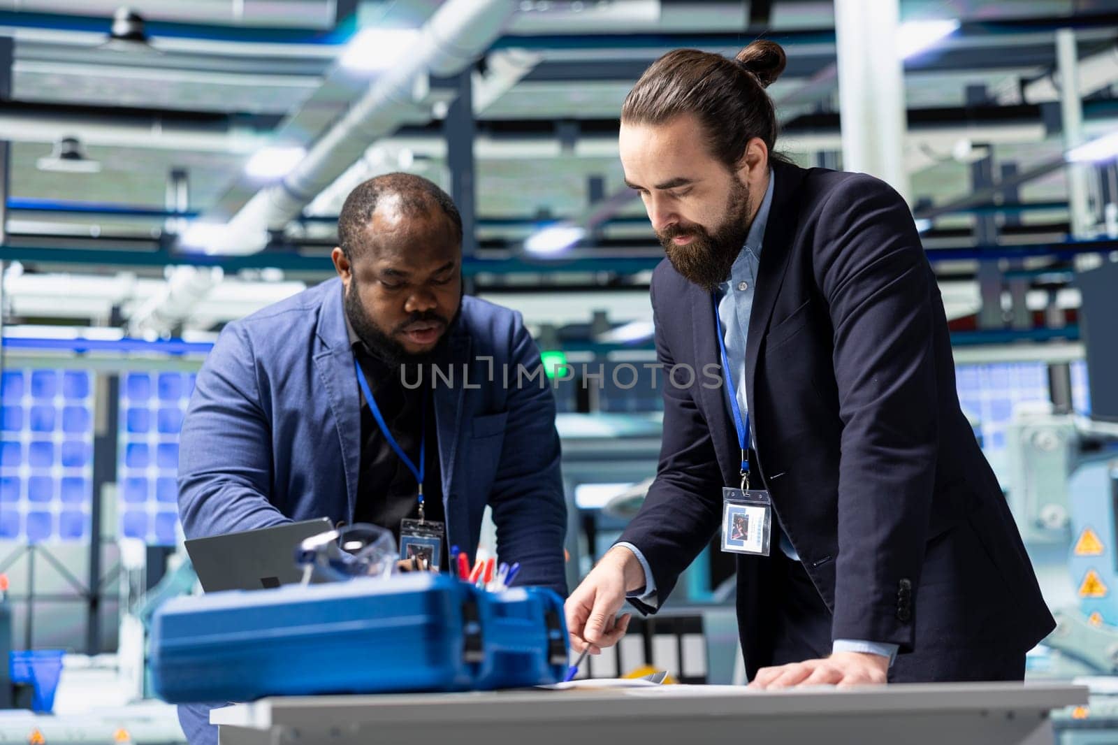 Multiethnic business associates and engineers gather for a discussion in a solar tech manufacturing facility, planning future developments in sustainable energy and industrial operations.