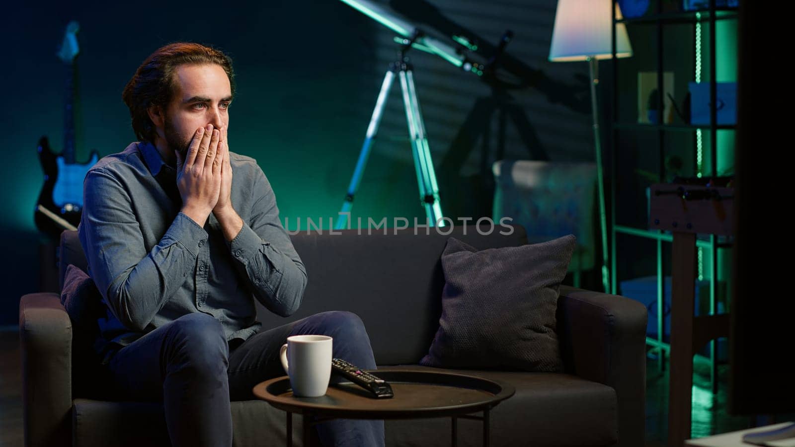 Man at home shocked by thriller film on TV, drinking coffee. Stupefied person at home left with eyes wide open by dramatic plot twist, watching suspenseful movie on TV, enjoying beverage, camera A