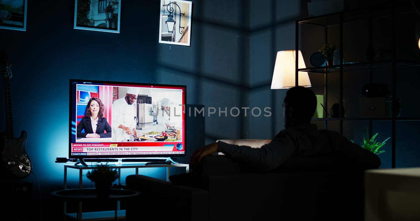 At night, young guy follows latest headlines on tv relaxing on couch in the living room, watching news program. On international network person pays attention to information broadcasted. Camera A.