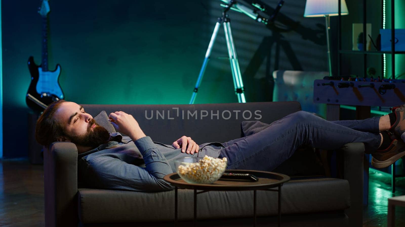 Smiling man lounged on couch at home eating popcorn while browsing though movies available on streaming service, enjoying time off from work. Man laying down in apartment consuming snacks, camera A