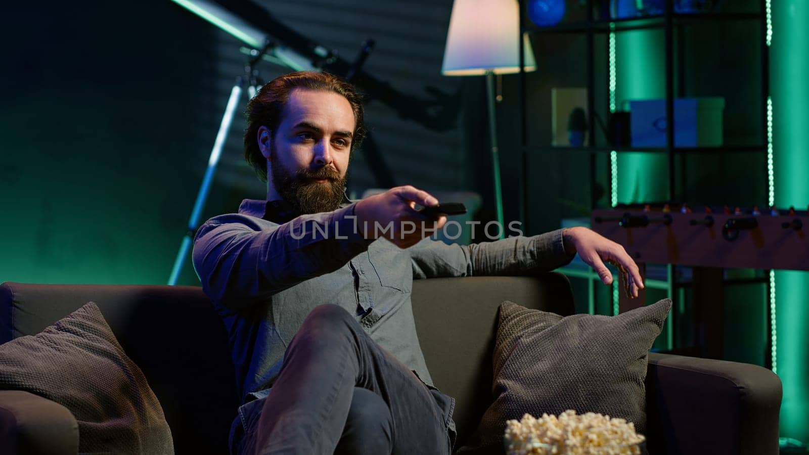 Man taking seat on sofa to watch television, getting entertained in relaxed home environment. Person arriving in apartment sitting down on couch to enjoy movies, turning on TV, eats popcorn, camera A