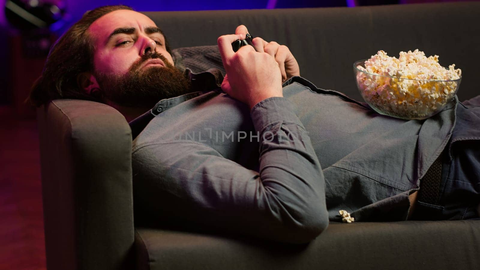 Gamer lounged on couch engaging in multiplayer game using gamepad, eating snack. Person in home theatre playing videogames on gaming console with bowl of popcorn in lap, camera A