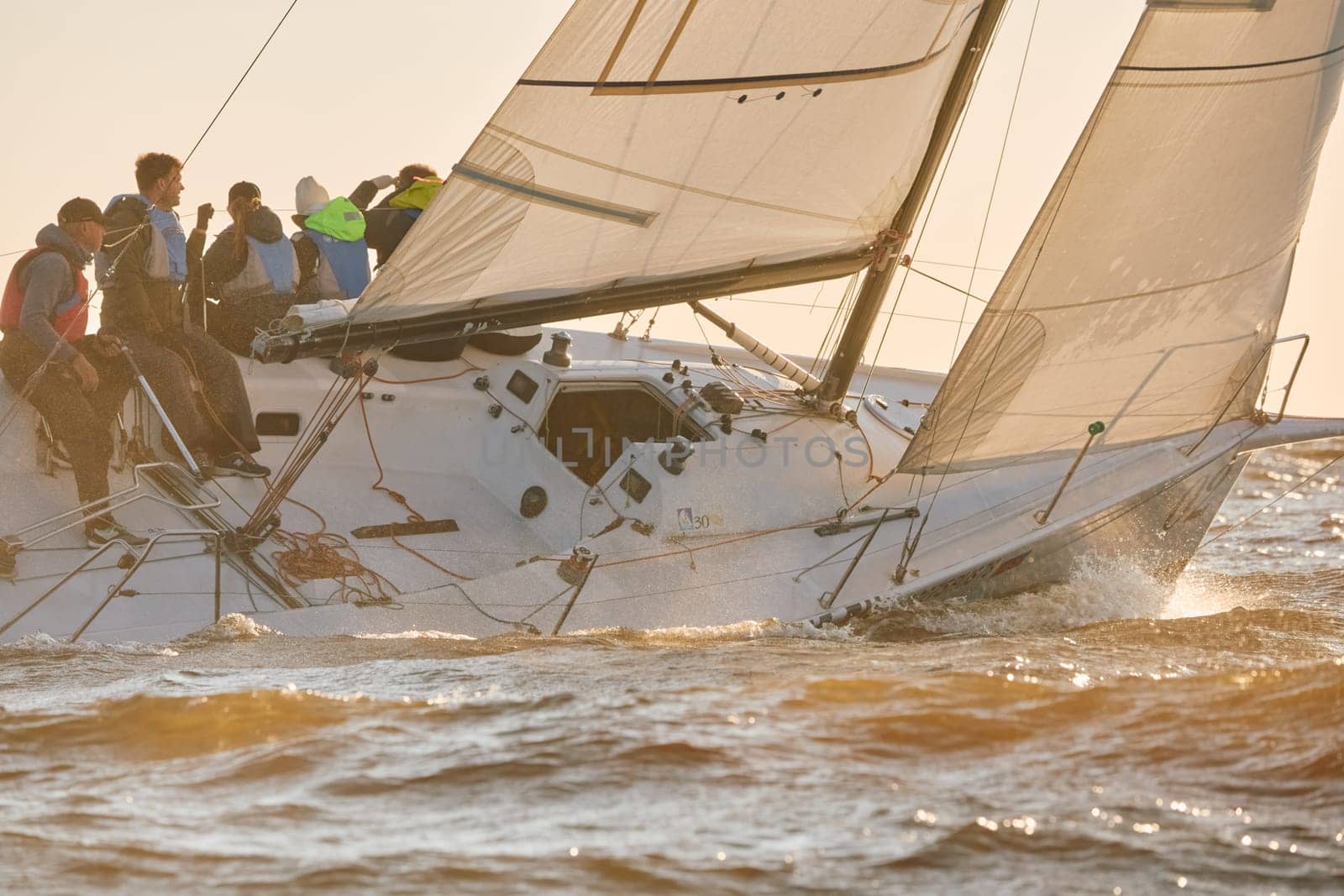 Russia, St.Petersburg, 07 June 2024: Sailing regatta in the Gulf of Finland at sunset, hot race of several yachts, teamwork, yachtsmen sitting on board the boat. High quality photo