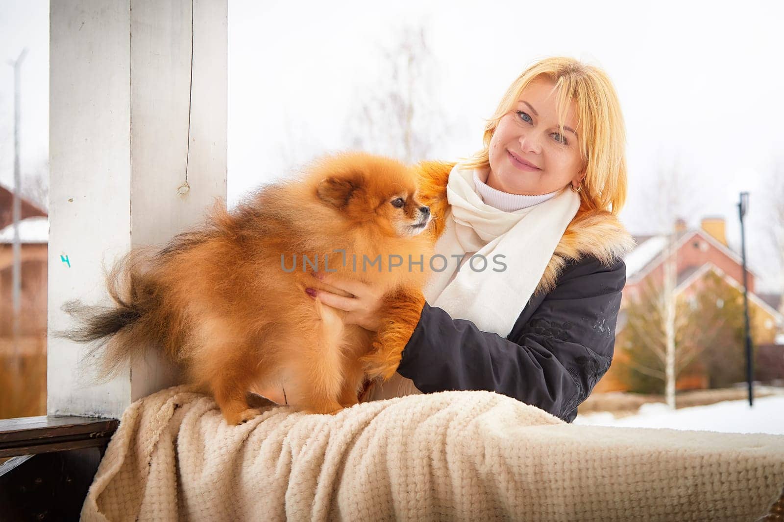 A fluffy red Pomeranian dog is held by its mature blonde owner, both enjoying a winter day in the snow.