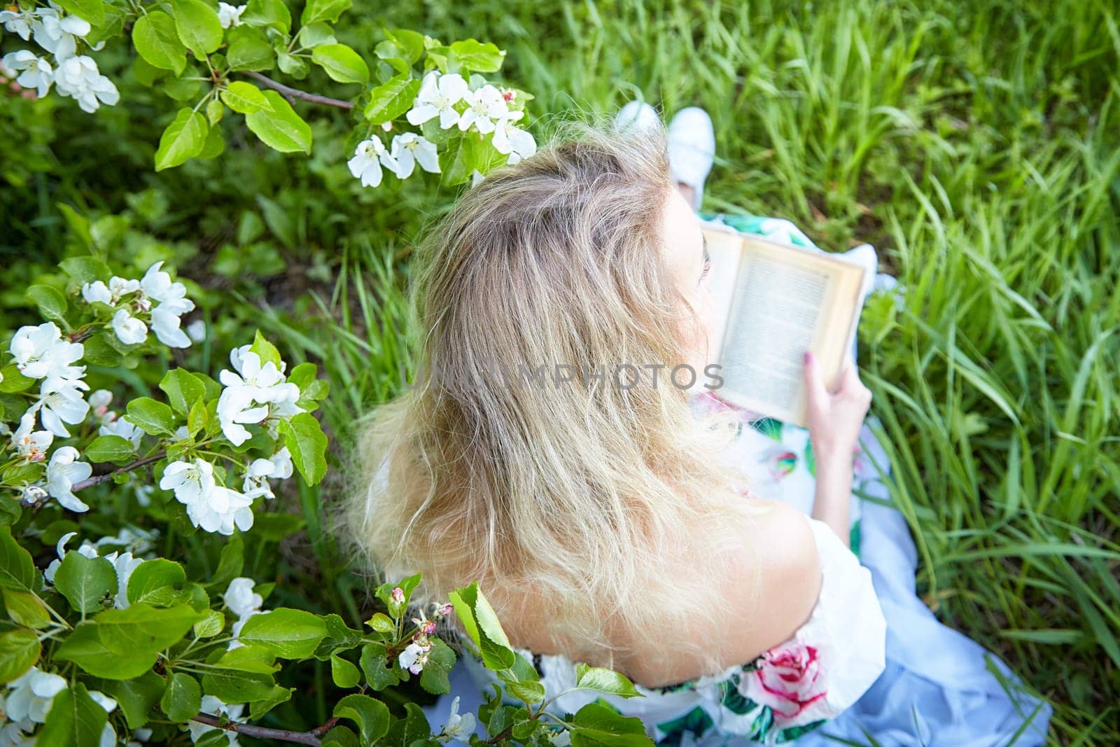 Mature woman with blonde hair sits in a lush garden, reading a book surrounded by vibrant flowers and grass on a sunny day.
