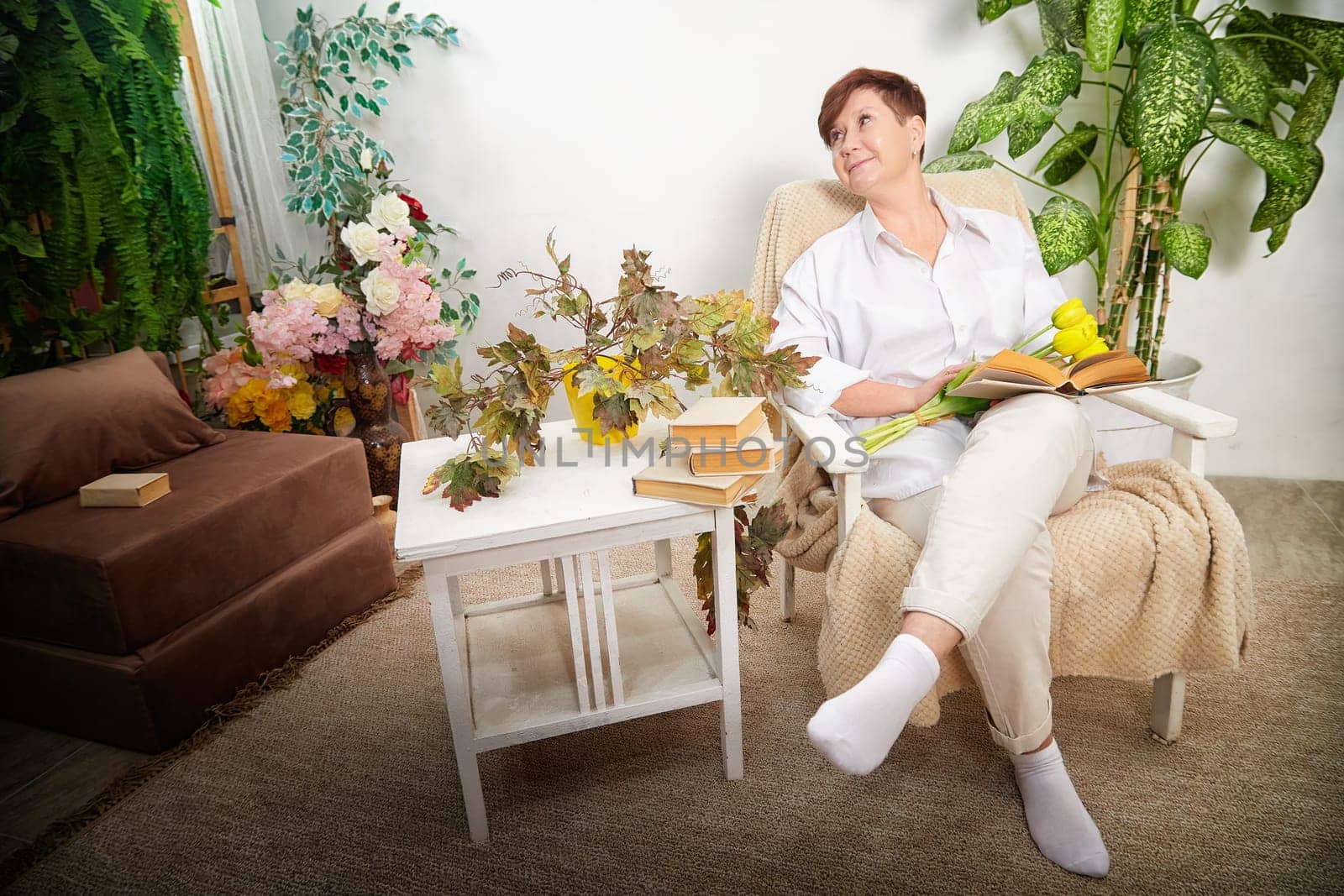 A middle-aged woman relaxes in a cozy room, surrounded by flowers and enjoying a book on International Women's Day.