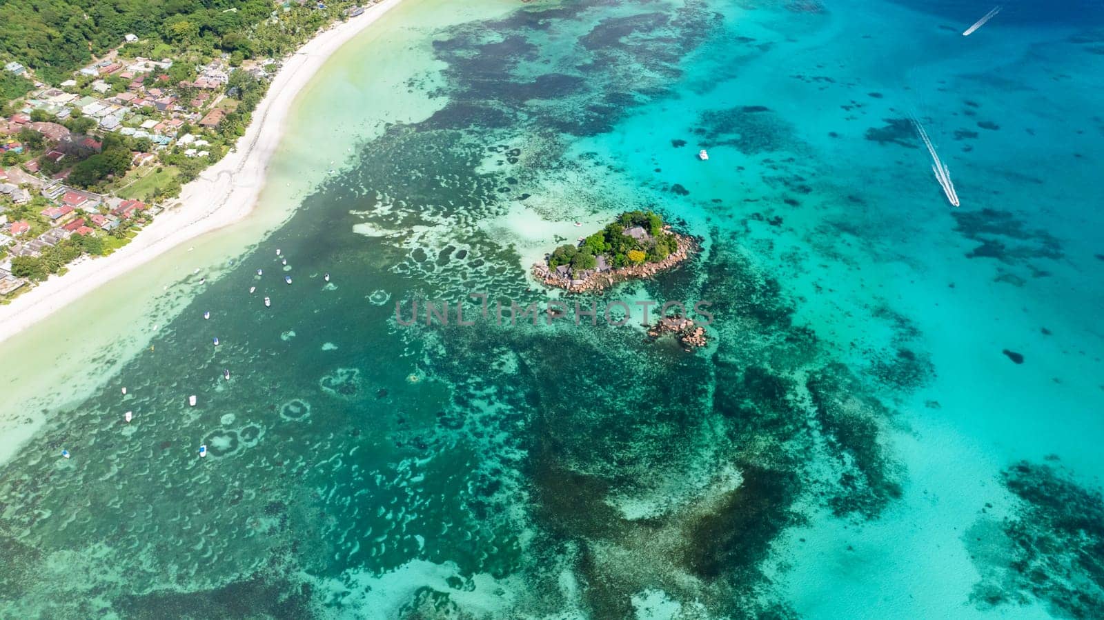 A small, lush island sits in shallow turquoise waters, surrounded by coral reefs and close to a white sandy shore. Praslin, Seychelles.