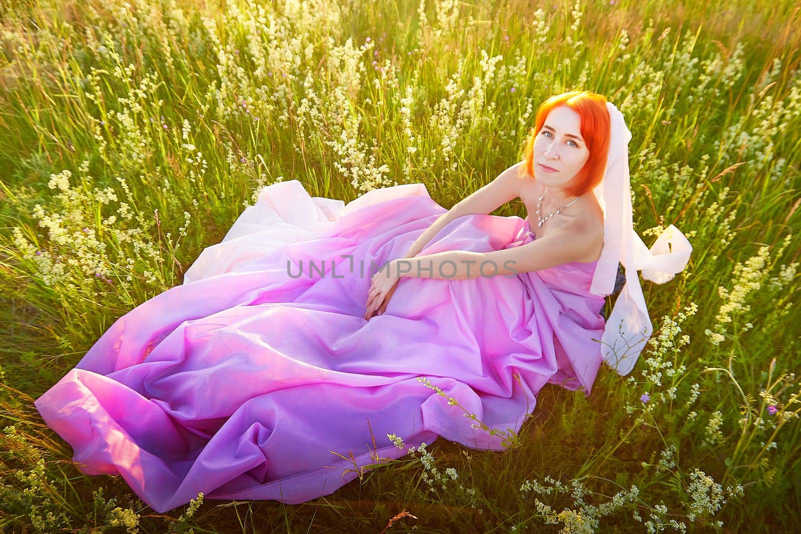 A model with vibrant orange hair sits gracefully on a blanket of pink fabric in a sunlit grassy field. Wildflowers surround her, creating a serene atmosphere at dusk.