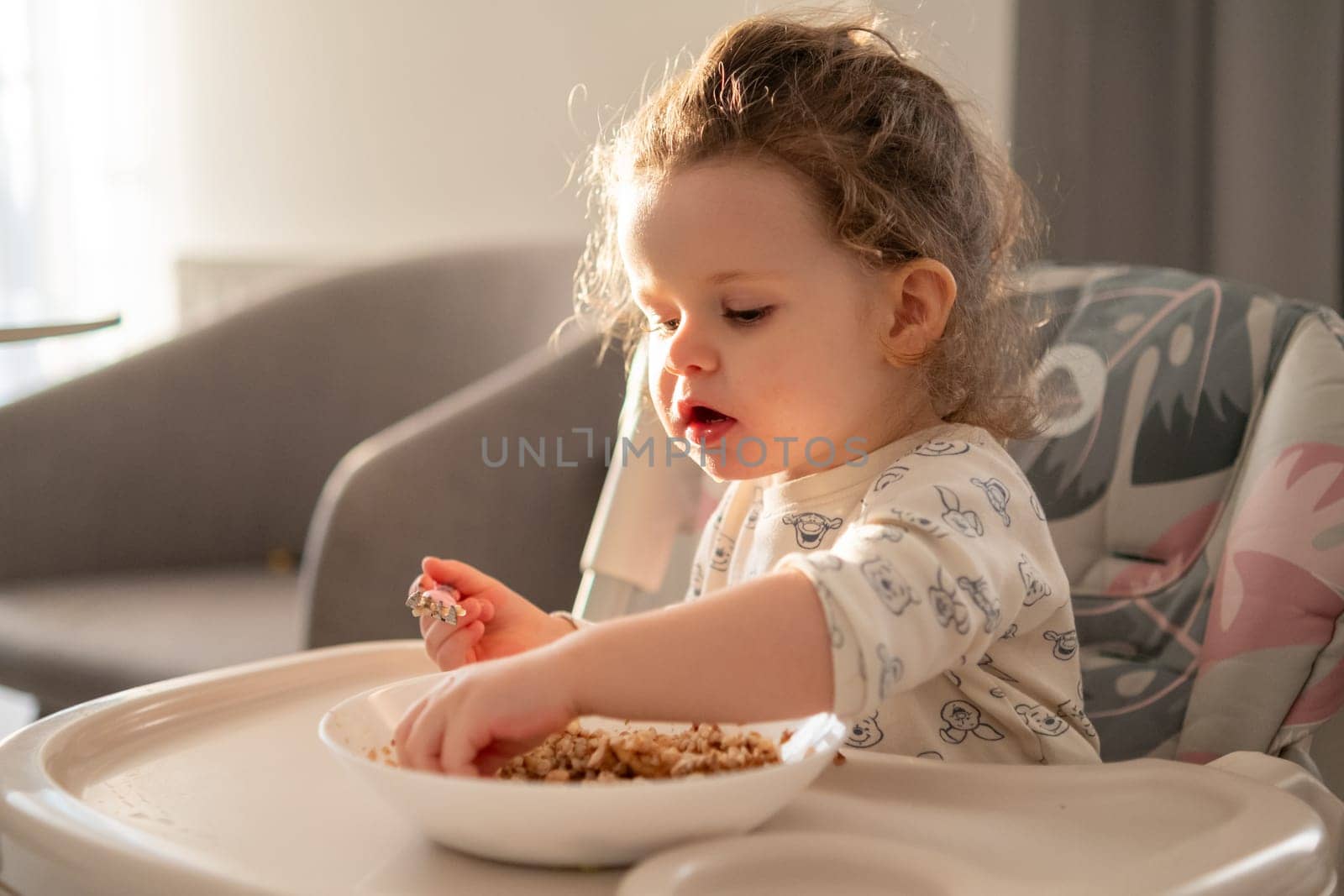 Small curly girl eating cereal at the table in a childs high chair. Natural light and warm home atmosphere.