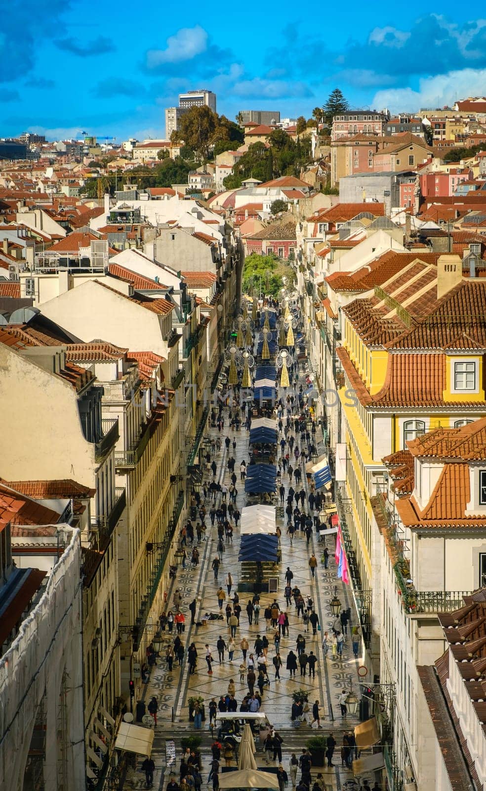 View of Rua Augusta in downtown Lisbon, Portugal, seen from above, crowds walking along iconic tiled pedestrian street, historic buildings with red rooftops, vibrant city life, bright blue sky with clouds, street decorations, outdoor cafes, dining