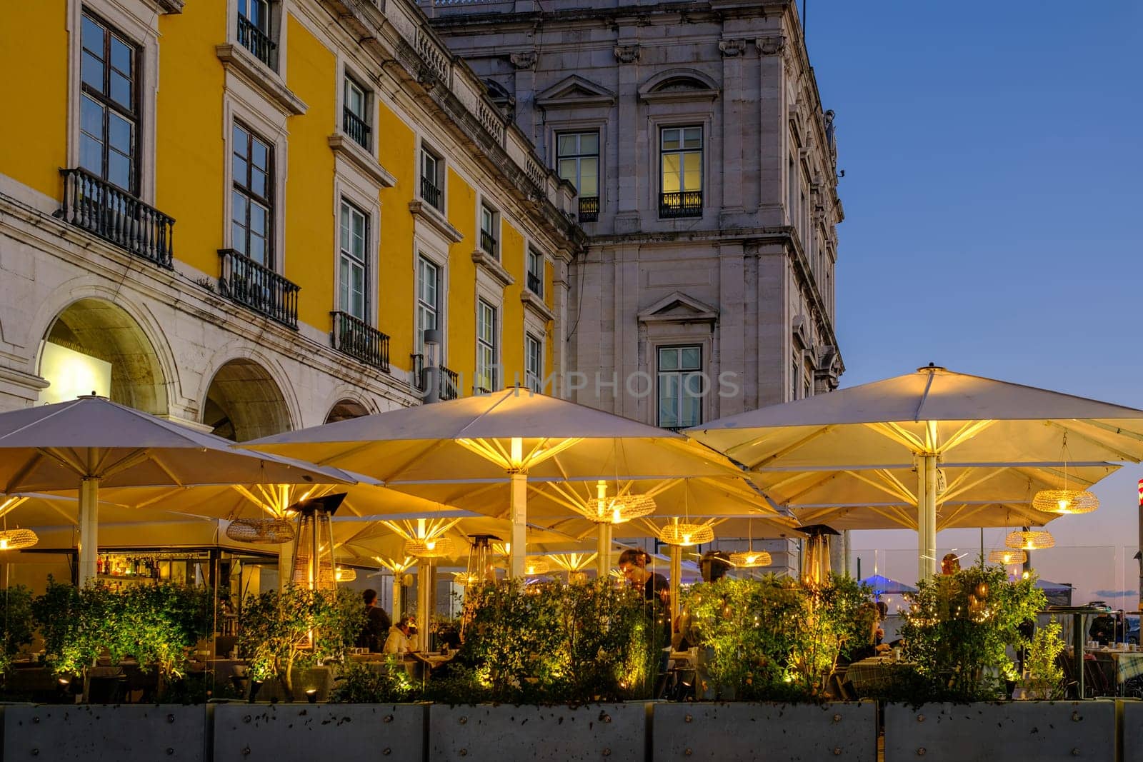 Elegant outdoor restaurant terrace in historic Commerce square, Lisbon, Portugal, with large illuminated umbrellas and lush greenery. People enjoy relaxed evening ambiance near iconic yellow buildings in city center, clear sky, sunset, twilight