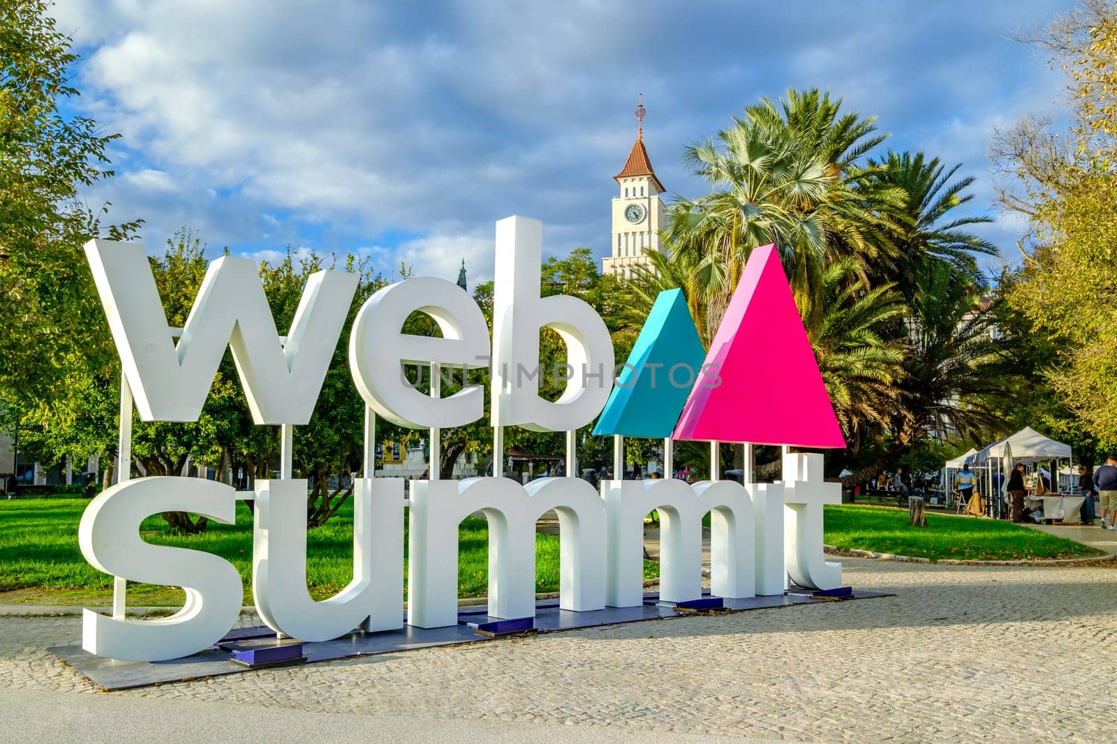 Nov 13, 2024 - Lisbon, Portugal: Colorful letters spell out Web Summit, influential technology conference, near park with lush palm trees and historic clock tower on sunny day. Discussions, network, innovative ideas, dynamic atmosphere of event