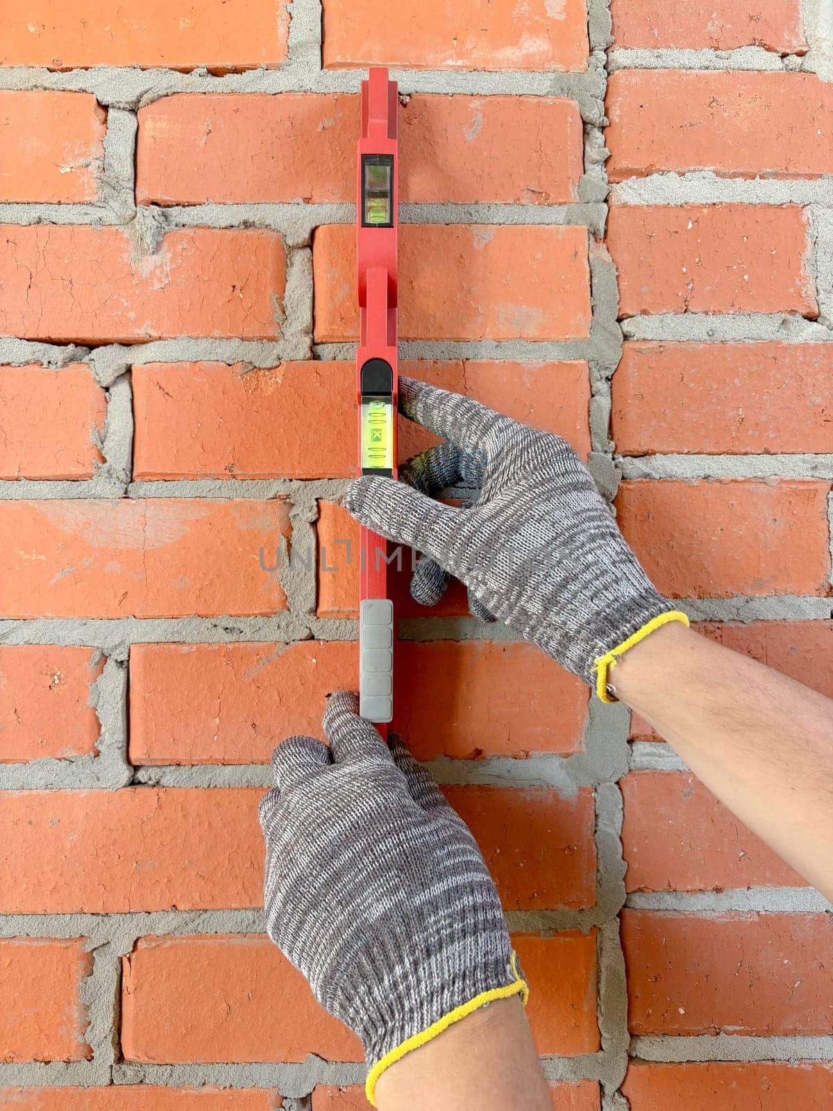A builder uses a bubble level to check a new red brick wall indoors. Construction and renovation of buildings. Vertical photo with hands and level on the wall.