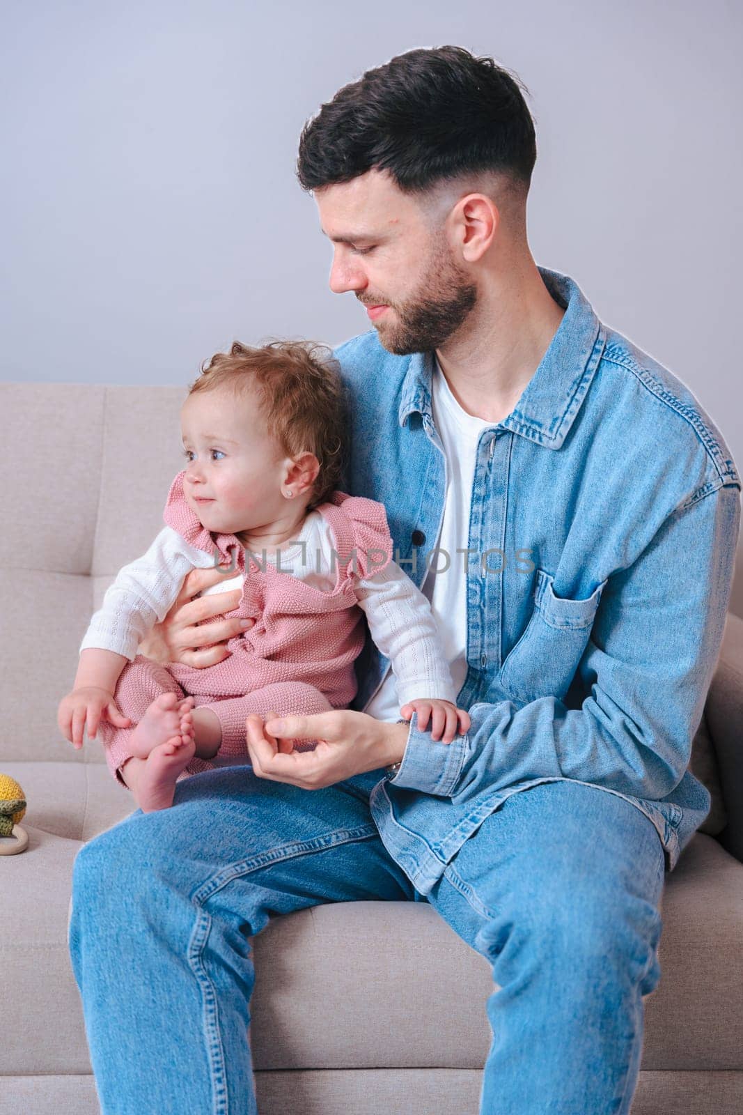Vertical portrait Young father in denim shirt gently holding his baby daughter while sitting on a sofa. The baby looks away curiously, capturing a calm and intimate moment of parenting at home.