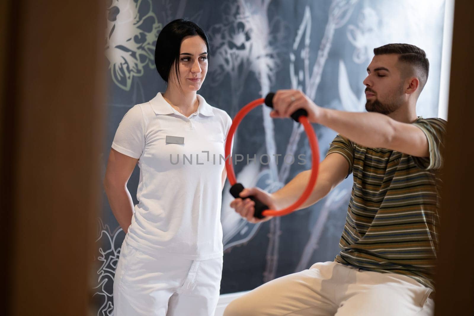 A male patient performing arm exercises using a resistance ring under supervision during a rehabilitation session.