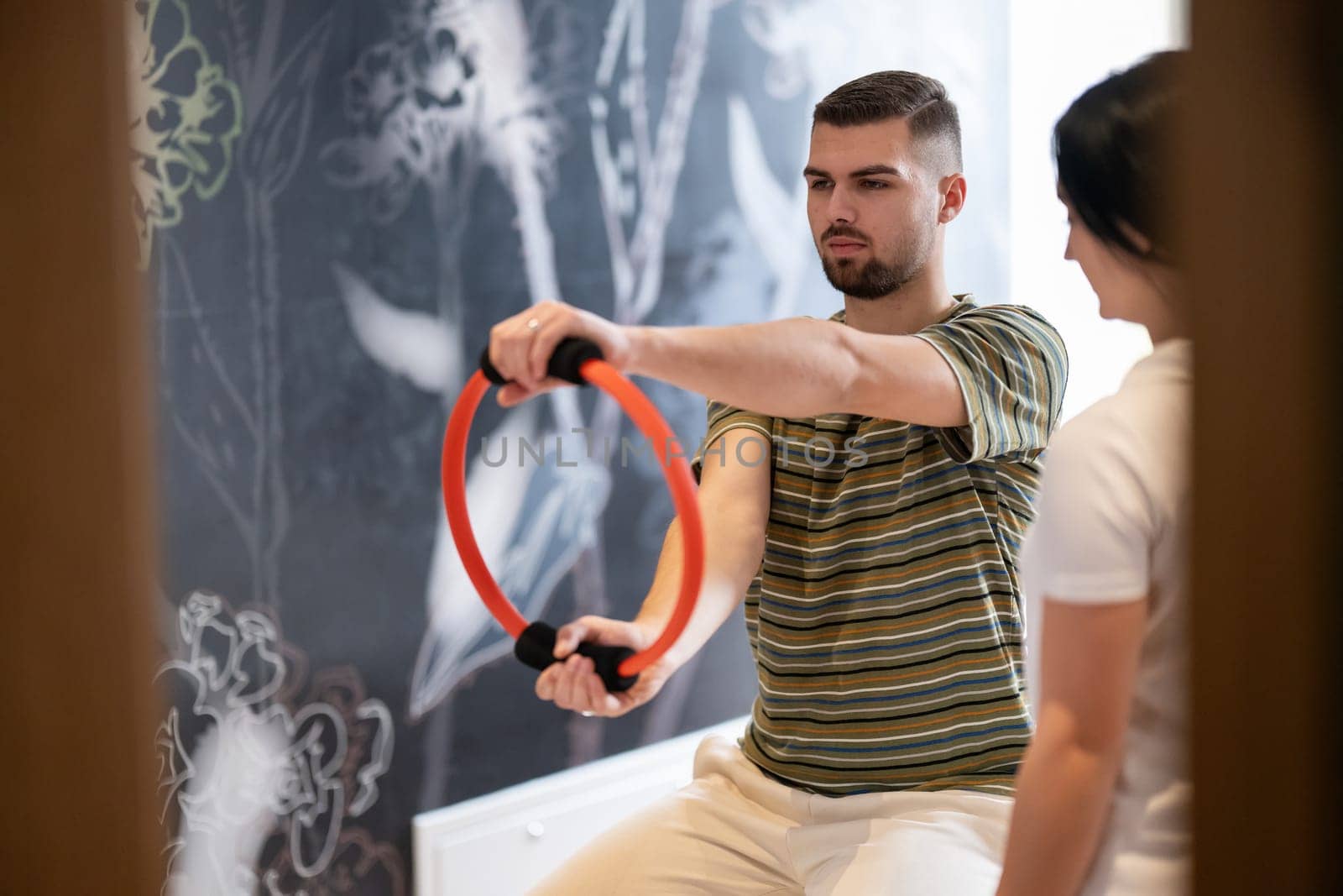 A male patient performing arm exercises using a resistance ring under supervision during a rehabilitation session.
