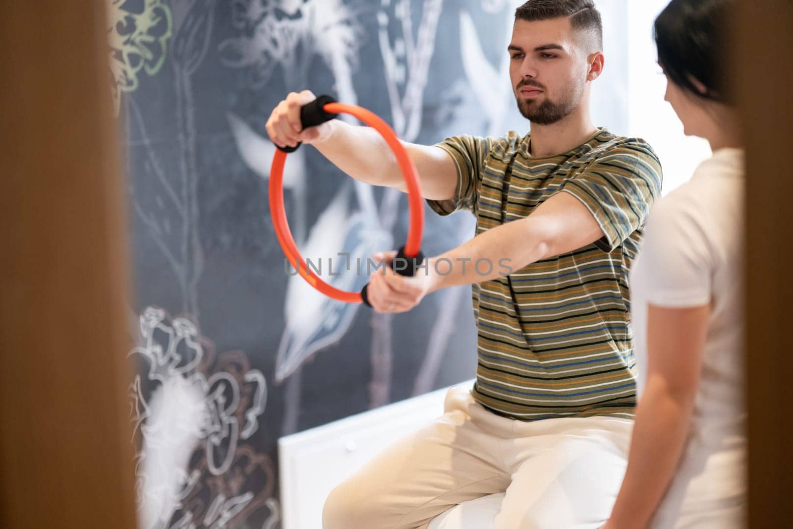 A male patient performing arm exercises using a resistance ring under supervision during a rehabilitation session.