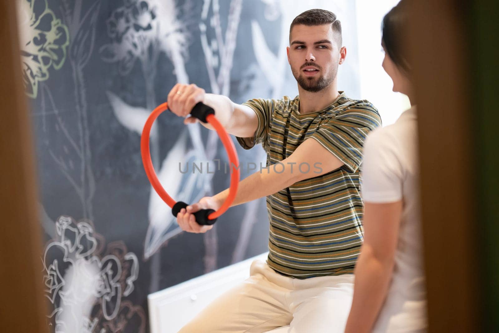 A male patient performing arm exercises using a resistance ring under supervision during a rehabilitation session.