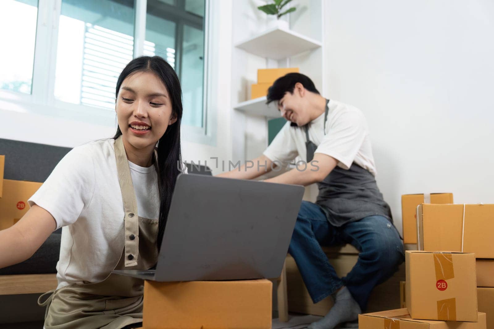 A couple shares a light moment while working on a laptop and handling packages, showcasing their partnership in a business setting.