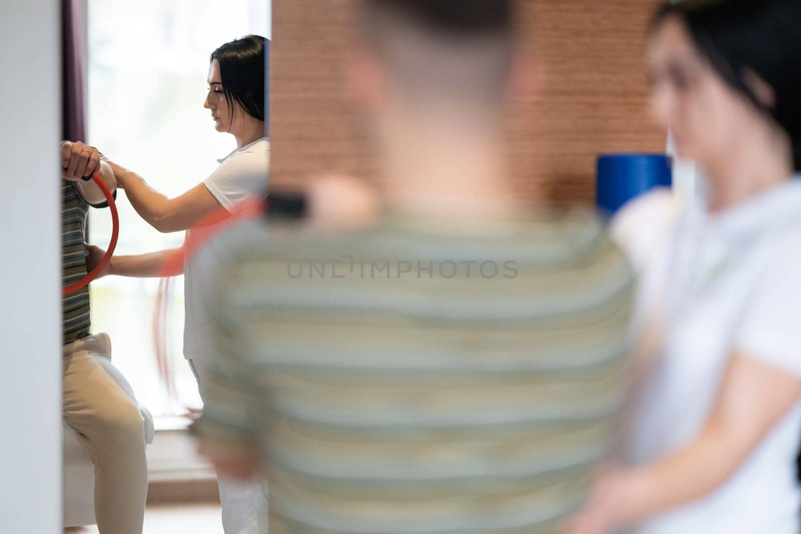 A male patient performing arm exercises using a resistance ring under supervision during a rehabilitation session.