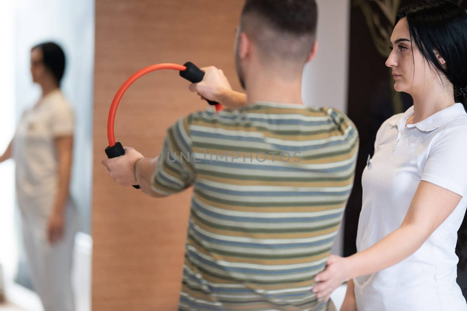 A male patient performing arm exercises using a resistance ring under supervision during a rehabilitation session.